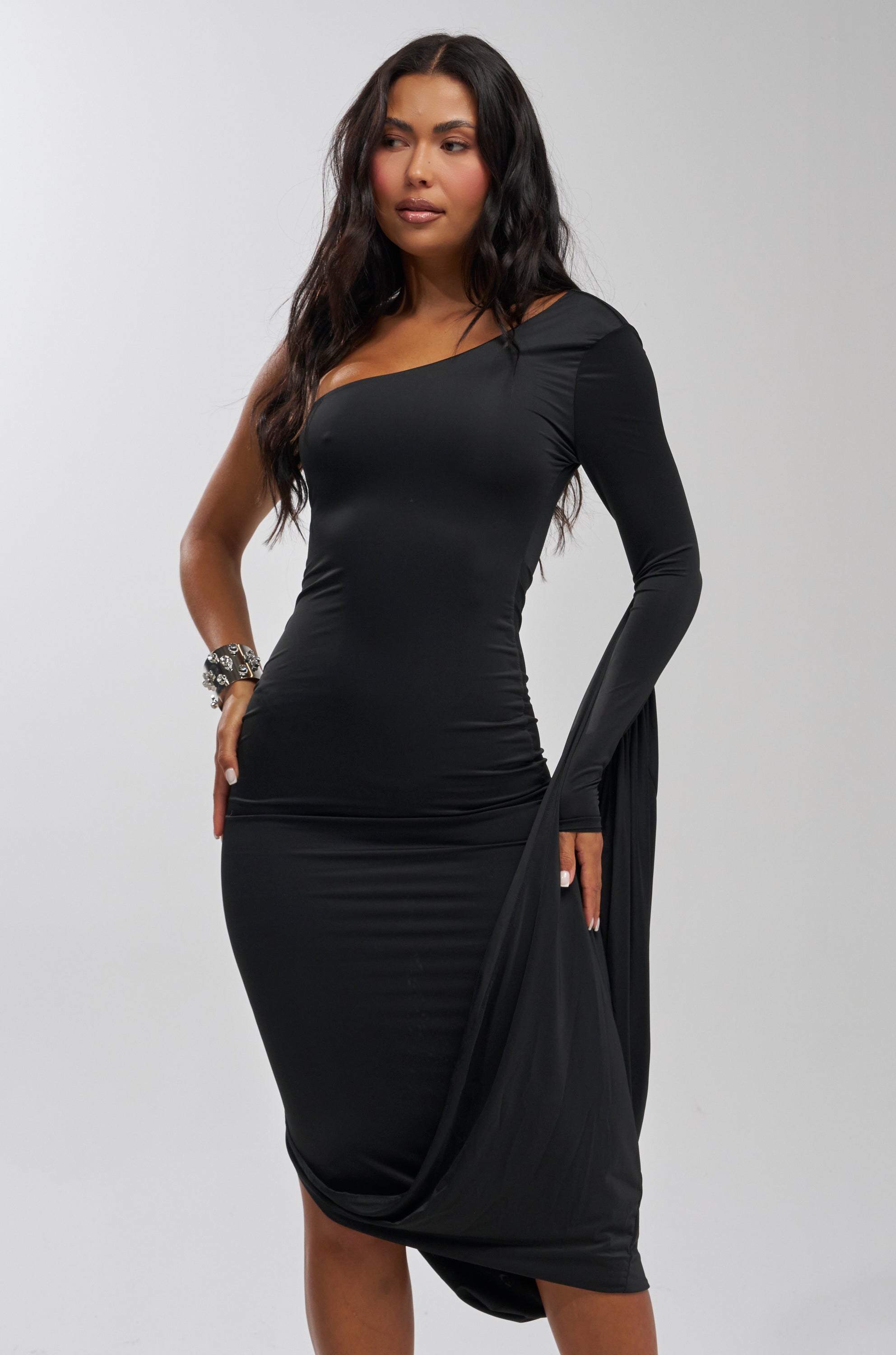 A woman poses confidently against a light background in the FOREVER FLIRTY ONE SHOULDER WINGED MIDI DRESS, a fitted black midi dress with an asymmetric neckline, draped winged silhouette, and one-shoulder design. She accessorizes with bracelets.