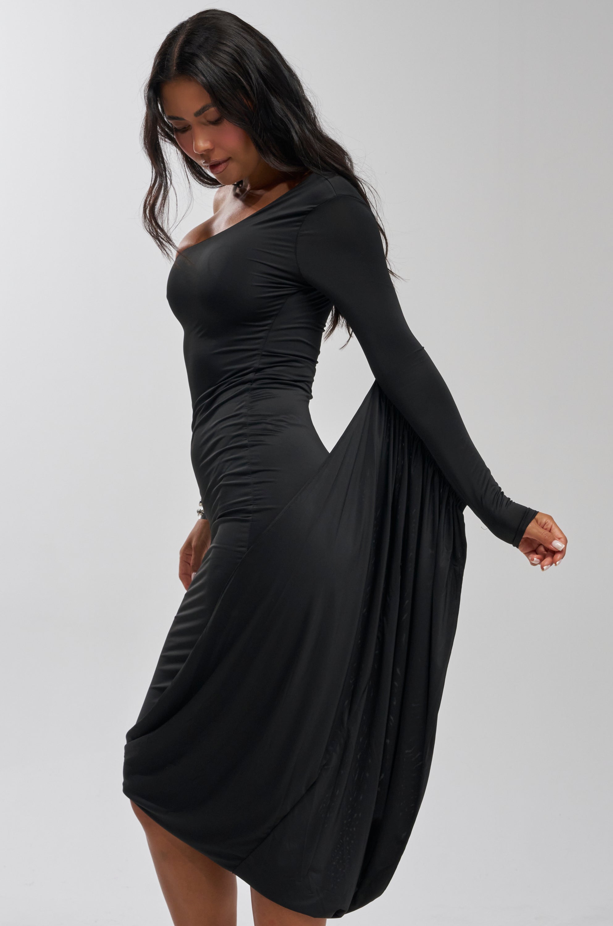 A woman models the FOREVER FLIRTY ONE SHOULDER WINGED MIDI DRESS, featuring a fitted black silhouette and dramatic draped wing, as she stands sideways against a plain light background with one arm extended behind her.