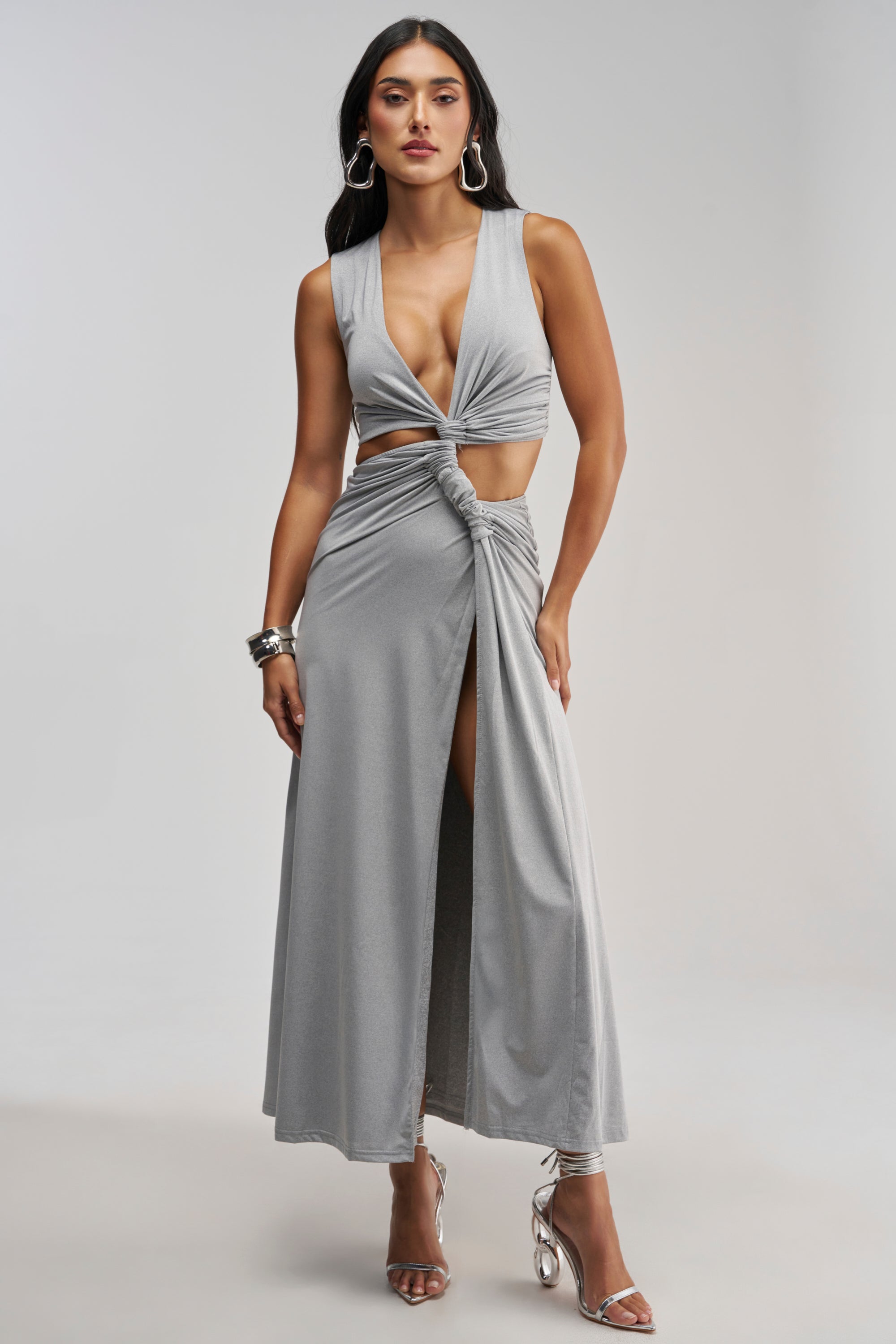 A woman poses against a plain background wearing the FIND ME IN THE PENTHOUSE MAXI DRESS IN HEATHER GREY—a sleeveless maxi dress featuring a knotted front, cutout detail, and thigh-high slit—paired with silver high heels. She accessorizes with large hoop earrings and several bangles, making this the perfect elegant evening choice for your next sexy vacation.