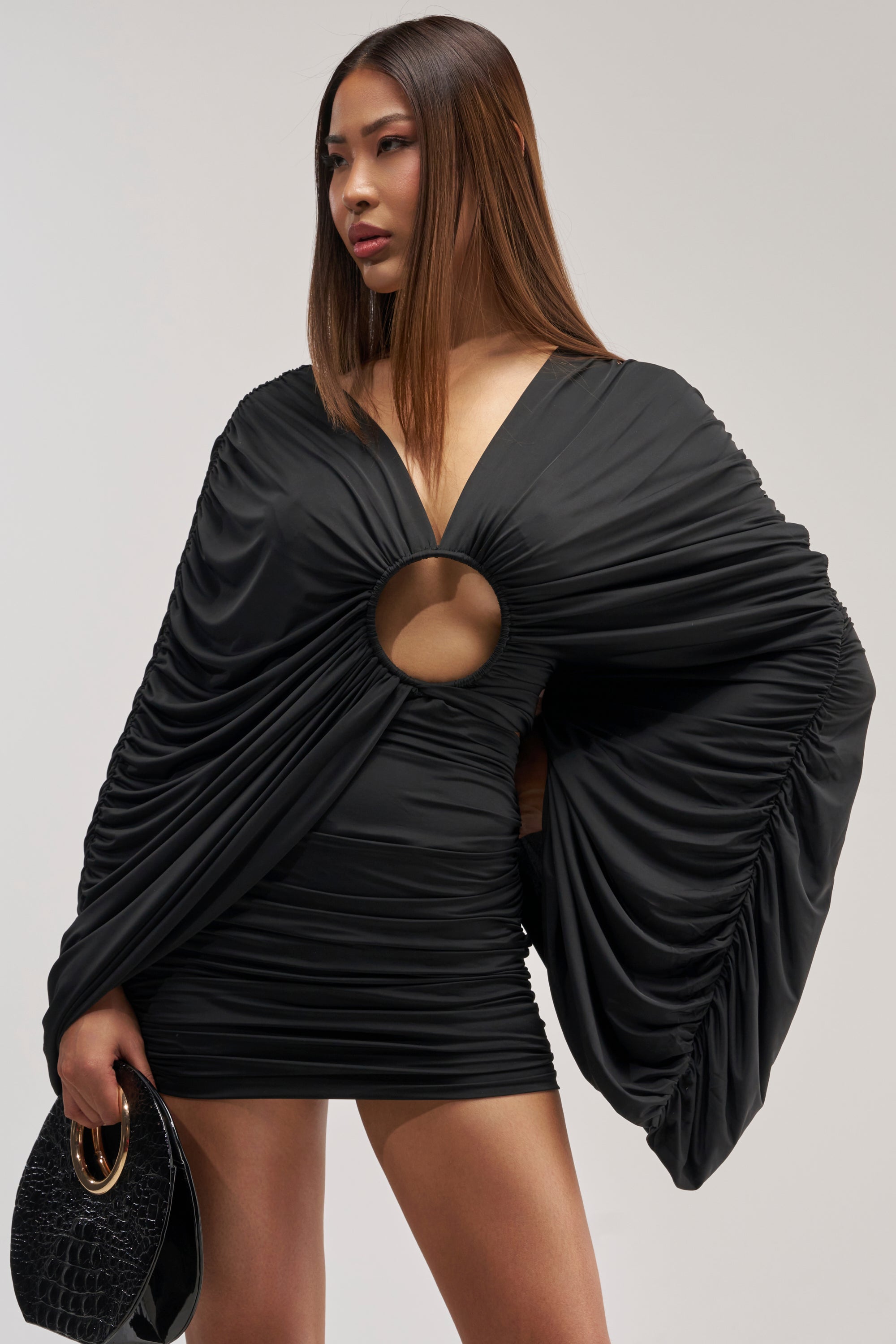 A woman models the BAD DECISIONS MINI DRESS IN BLACK, featuring dramatic ruched sleeves and a circular cutout at the front, while holding a black handbag and posing against a plain background.