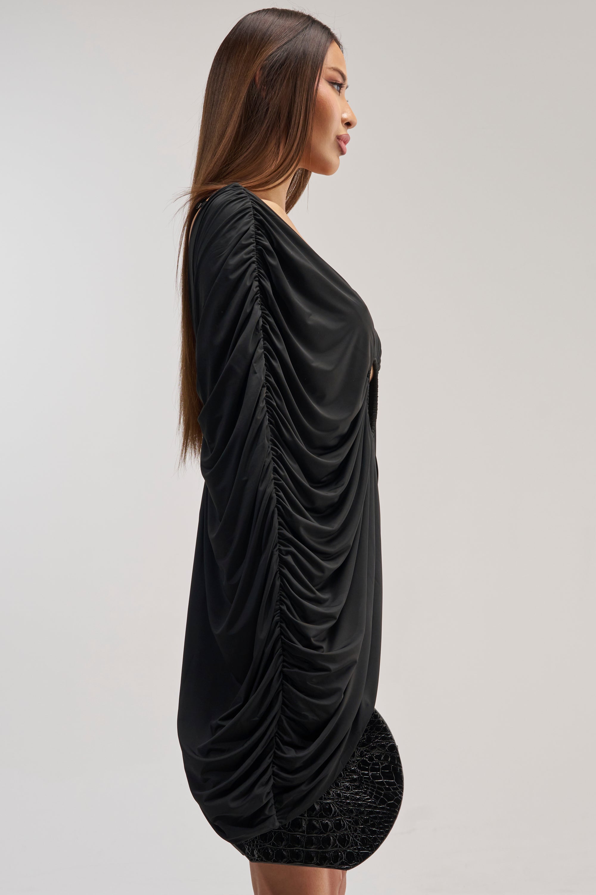 A woman in profile stands against a plain light gray background, wearing the BAD DECISIONS MINI DRESS IN BLACK—a loose, ruched black dress with long sleeves—her straight brown hair framing her face.