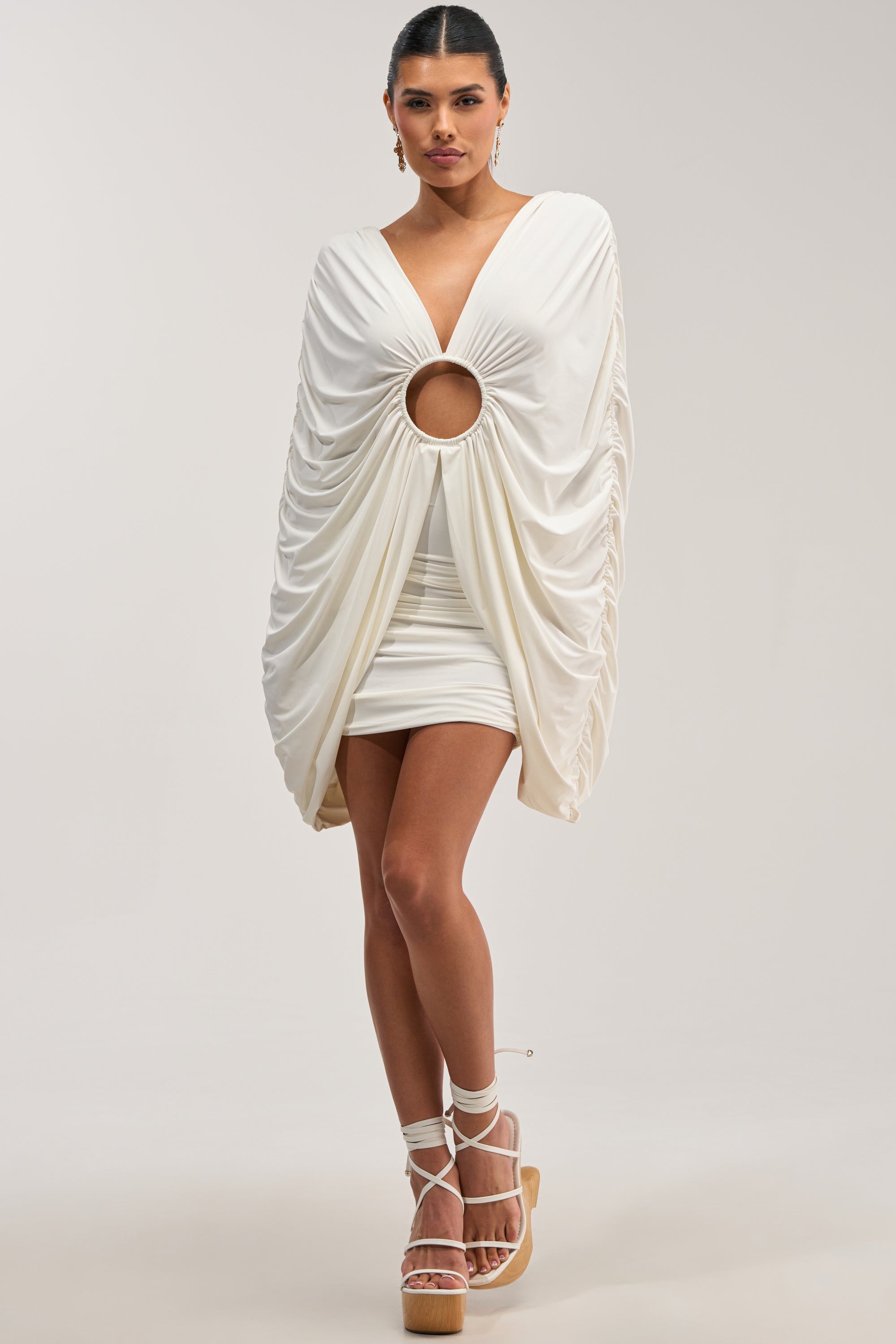 A woman poses in the BAD DECISIONS MINI DRESS IN WHITE, featuring dramatic draped sleeves, a deep V neckline, and a large circular cutout at the waist. She completes the look with platform sandals with ribbon ties and has her hair pulled back.