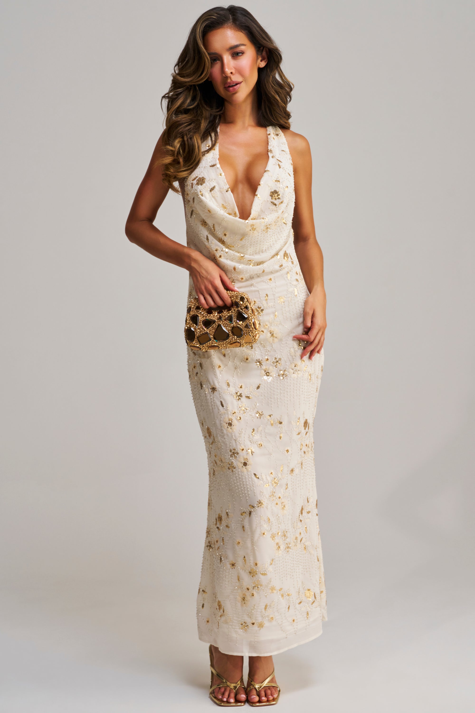 A woman models the MAKE ME BLUSH EMBELLISHED MAXI DRESS, a floral halter gown with gold accents. She accessorizes with a gold clutch and heels, her long wavy hair flowing as she poses against a plain light background.
