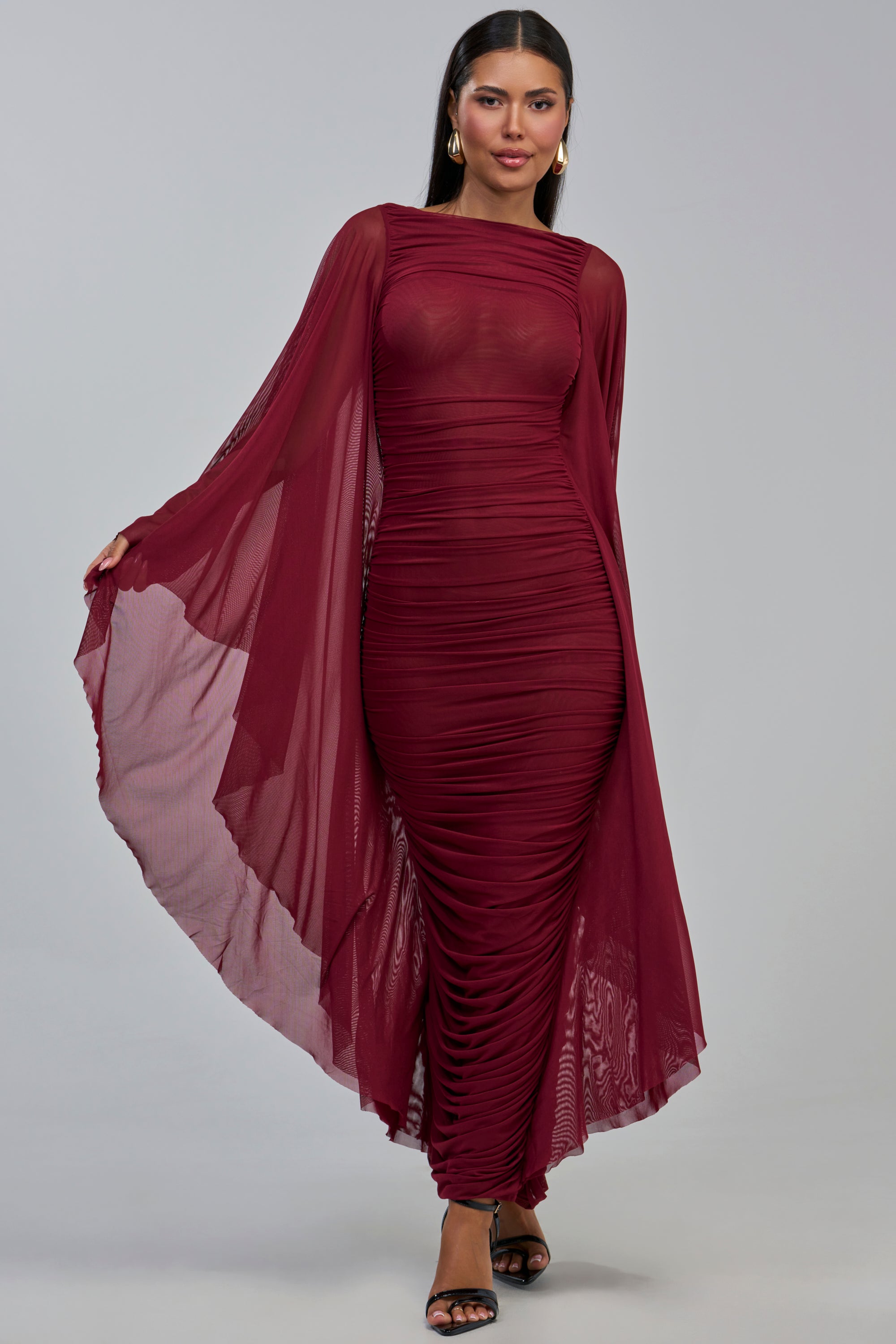 A woman models the SHE IS THE MOMENT MESH MAXI DRESS, a fitted, sheer burgundy mesh maxi dress featuring dramatic cape sleeves and ruched detailing. She pairs it with black heels while standing against a plain light gray background—an elegant option for an evening event or as a wedding guest dress.
