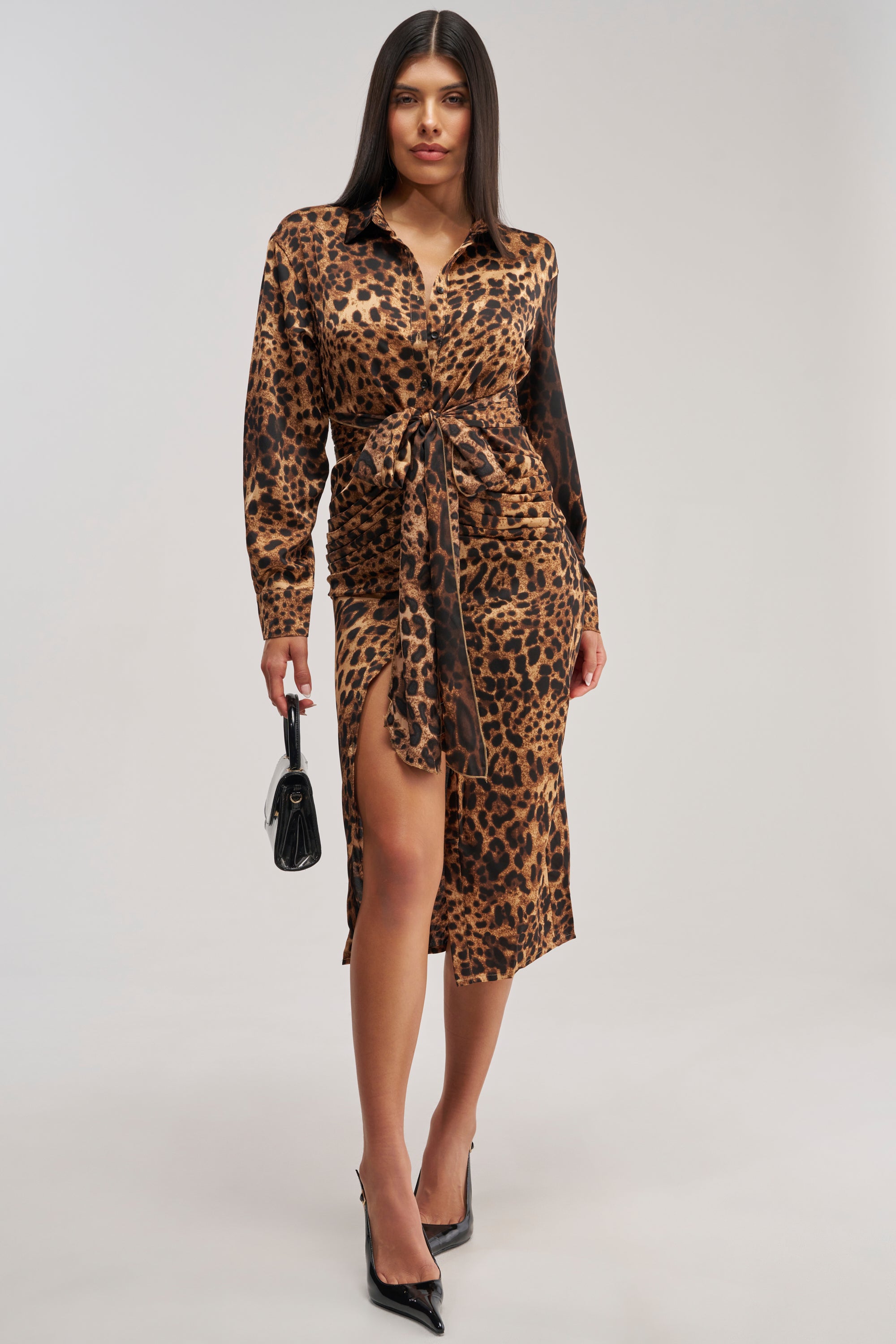The woman is wearing the GOLDEN RADIANCE CHEETAH MIDI DRESS, a long-sleeve, cheetah print midi dress with a front knot and side slit. She pairs it with black heels and a small black handbag, standing against a plain background.