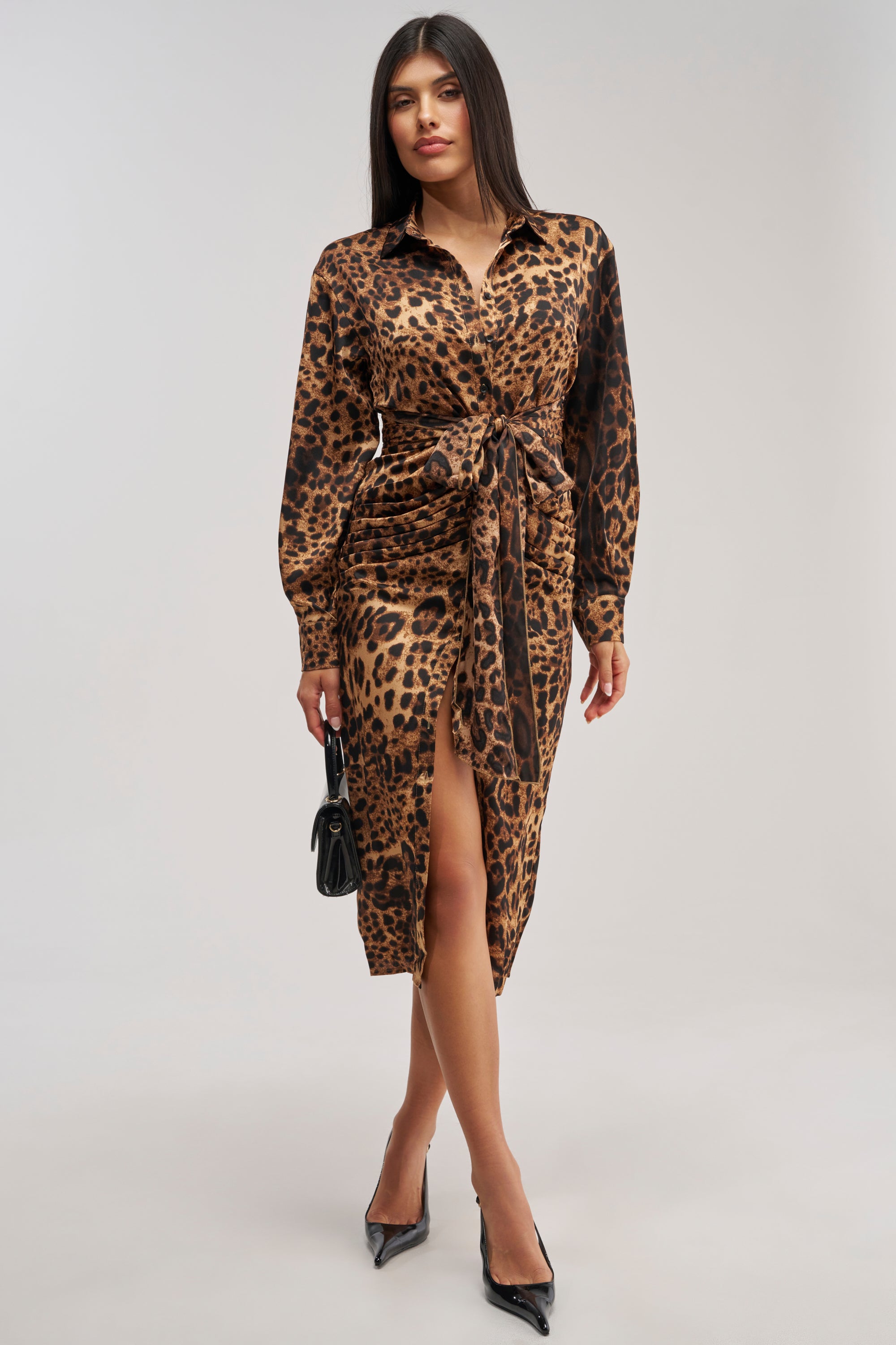 A woman poses against a plain light background wearing the GOLDEN RADIANCE CHEETAH MIDI DRESS, featuring long sleeves and a waist tie, paired with black pointed heels and a small black handbag.