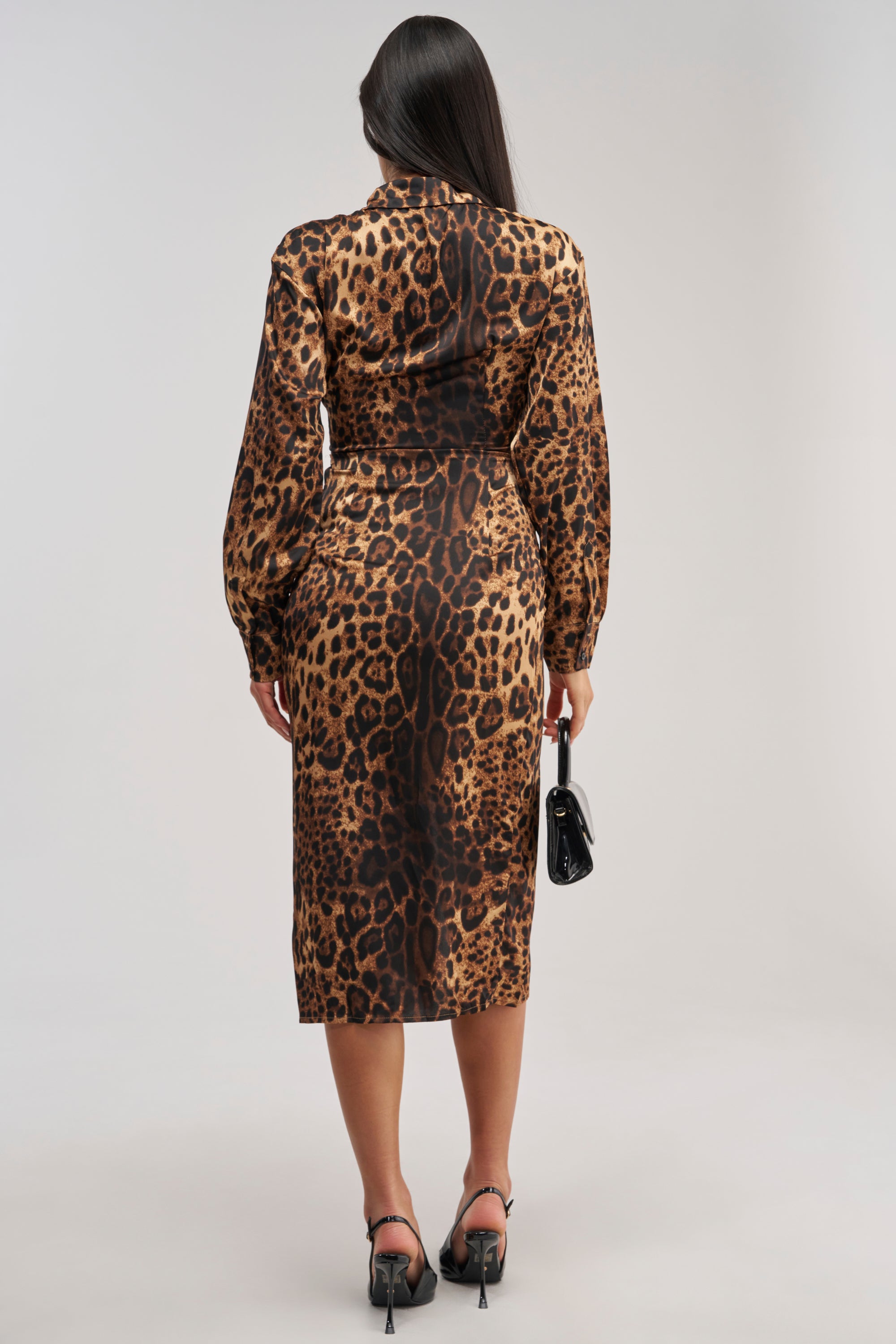 A woman stands facing away against a plain light background, wearing the GOLDEN RADIANCE CHEETAH MIDI DRESS with long sleeves, paired with black high-heeled shoes and holding a small black handbag.