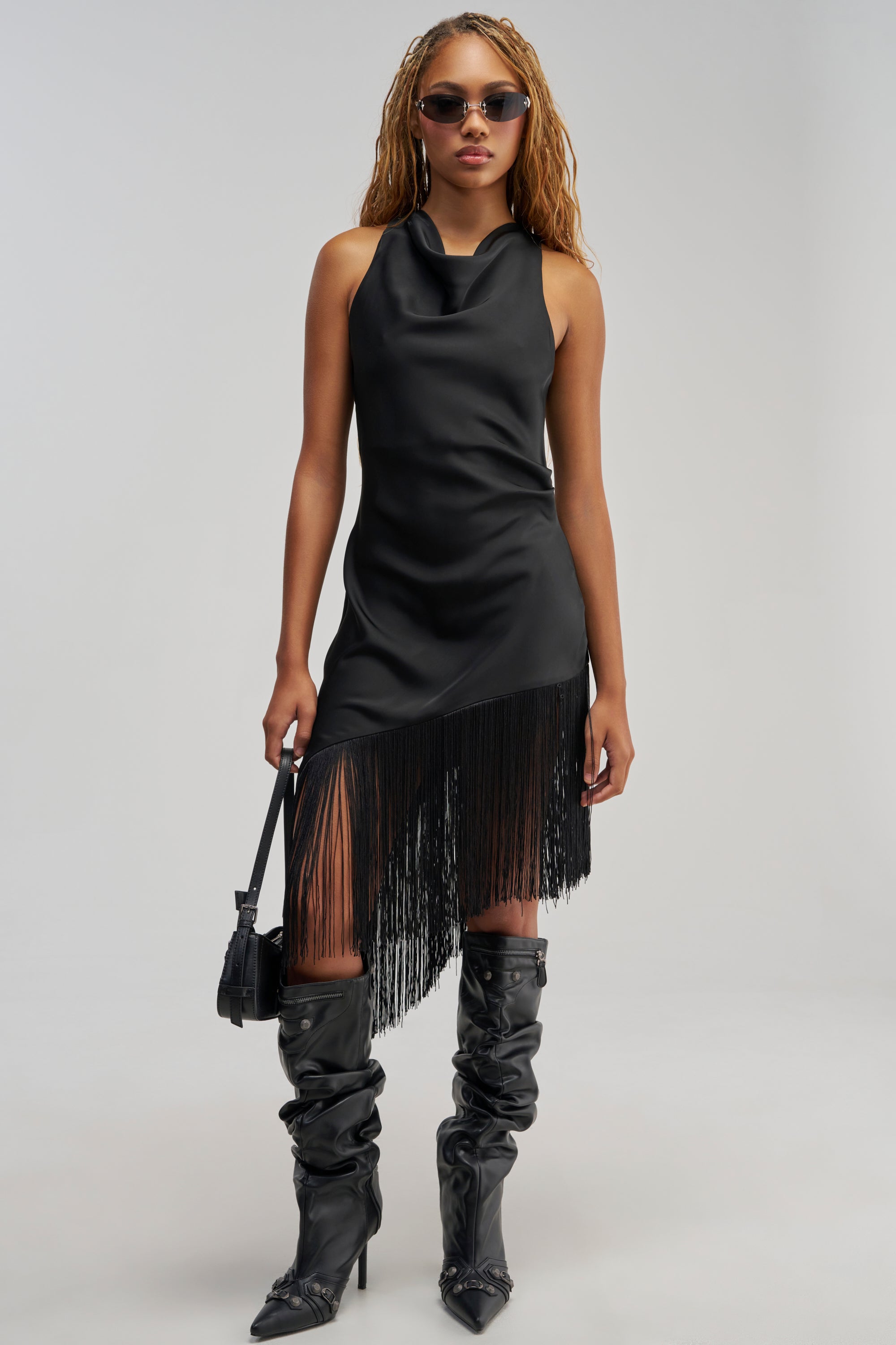 A woman in sunglasses wears the GRAND REVEAL FRINGE MIDI DRESS—a sleeveless black satin midi dress with fringe detailing at the hem—paired with black ruched knee-high boots and a small black purse, standing against a plain light background.