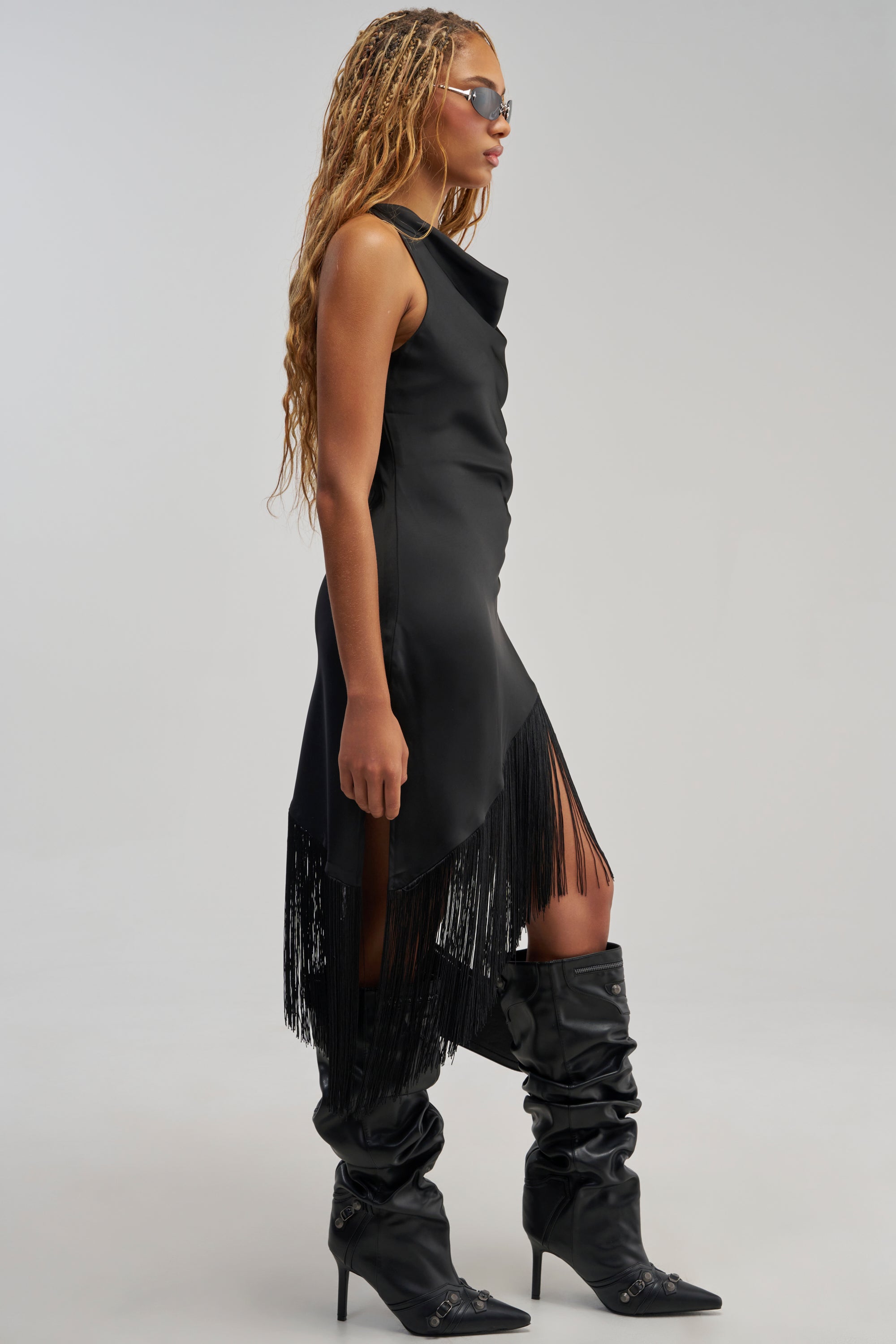 A person wearing sunglasses and the GRAND REVEAL FRINGE MIDI DRESS—a black satin, sleeveless midi dress featuring fringe detailing at the hem—paired with black slouchy heeled boots, stands in profile against a light gray background for an effortlessly chic look that’s perfect as an evening dress or stylish wedding guest dress.