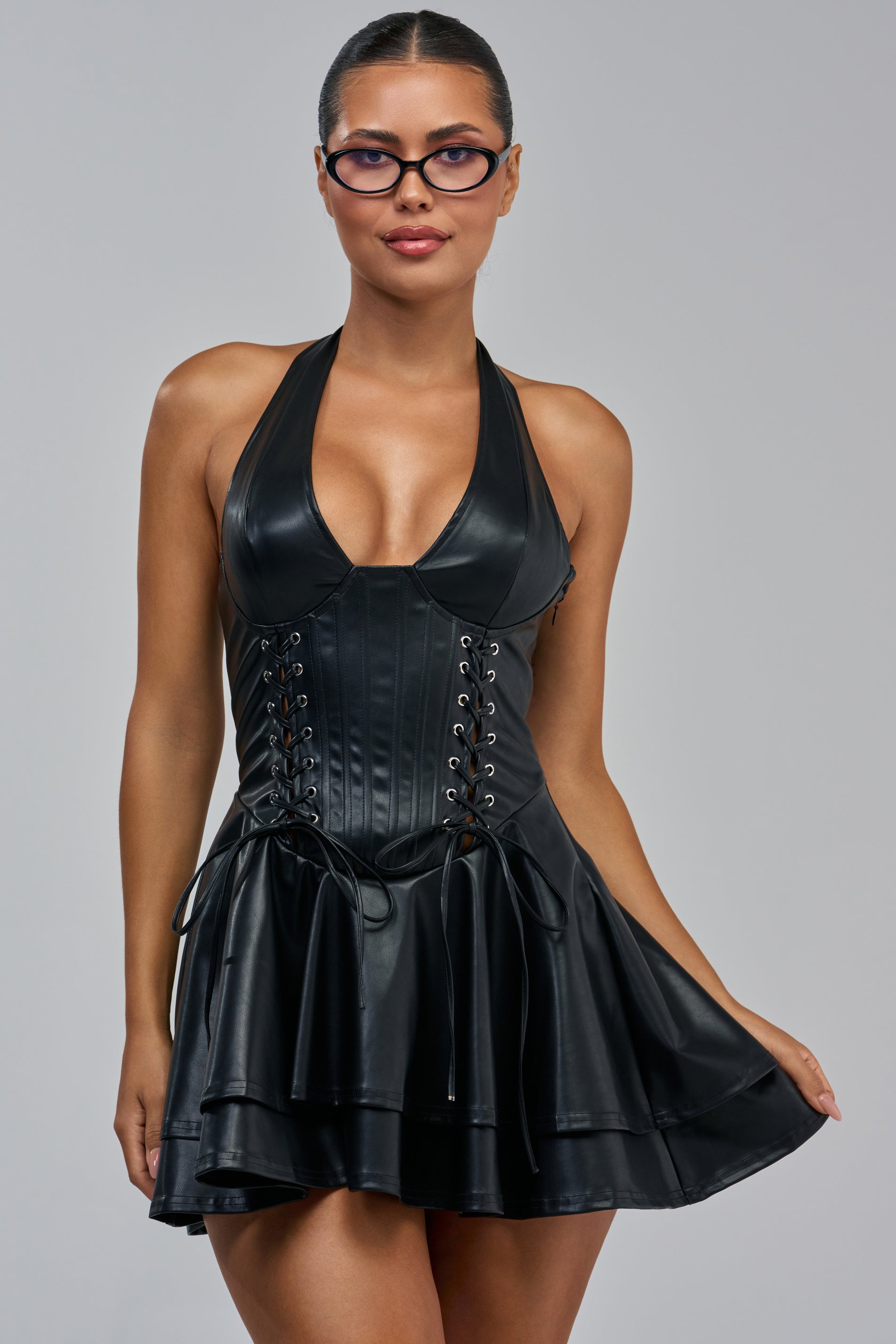 A woman wearing glasses models the VENOMOUS LACE UP PU MINI DRESS IN BLACK, featuring a halter-neck, lace-up corset detail, and a layered skirt. She poses against a plain gray background in this edgy yet elegantly detailed party dress.