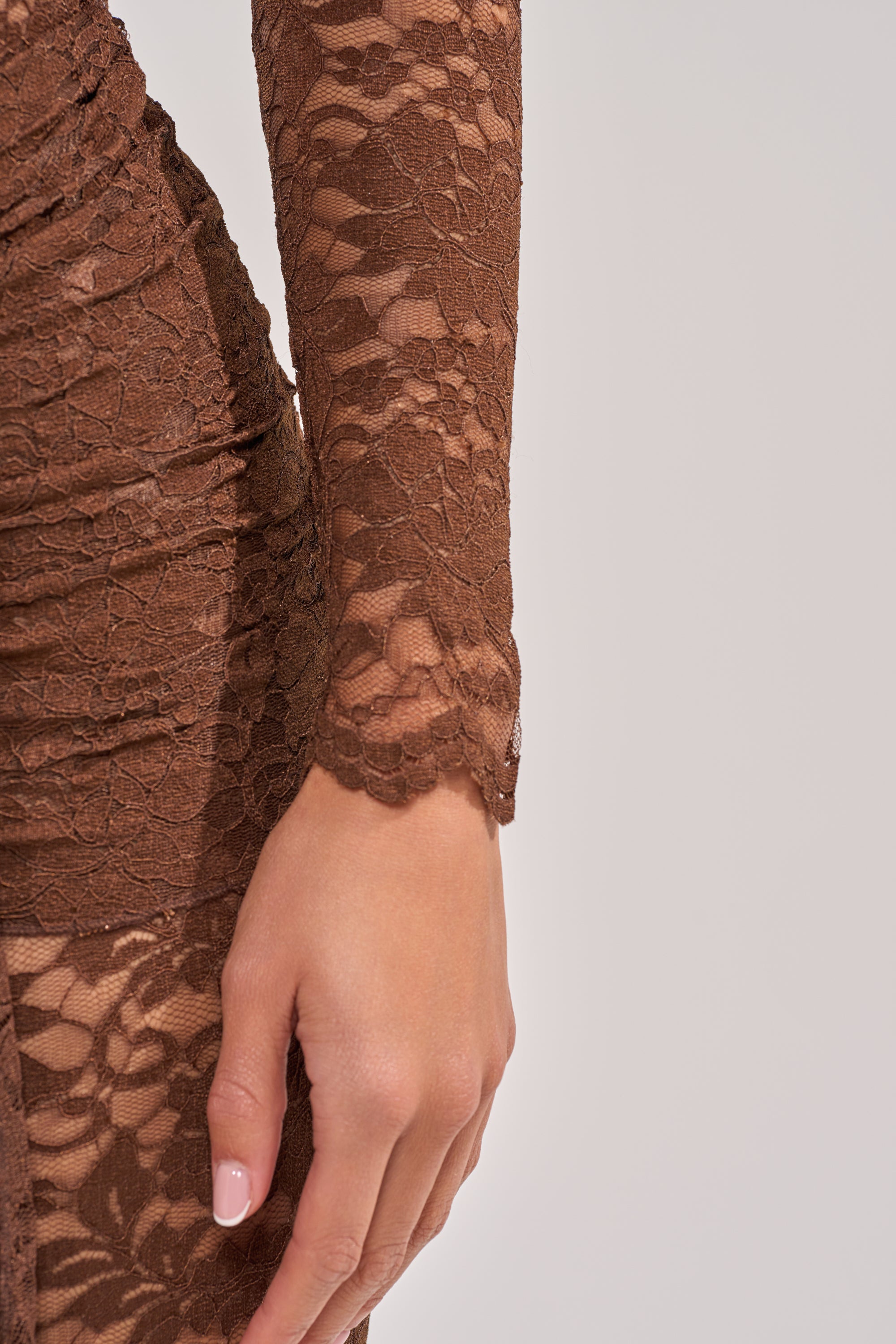 A close-up shows a hand resting by the side, wearing the ICONIC BEHAVIOR LACE CAPRI JUMPSUIT—a fitted brown lace piece with long sleeves and a subtle floral pattern—against a plain, light-colored background. 5