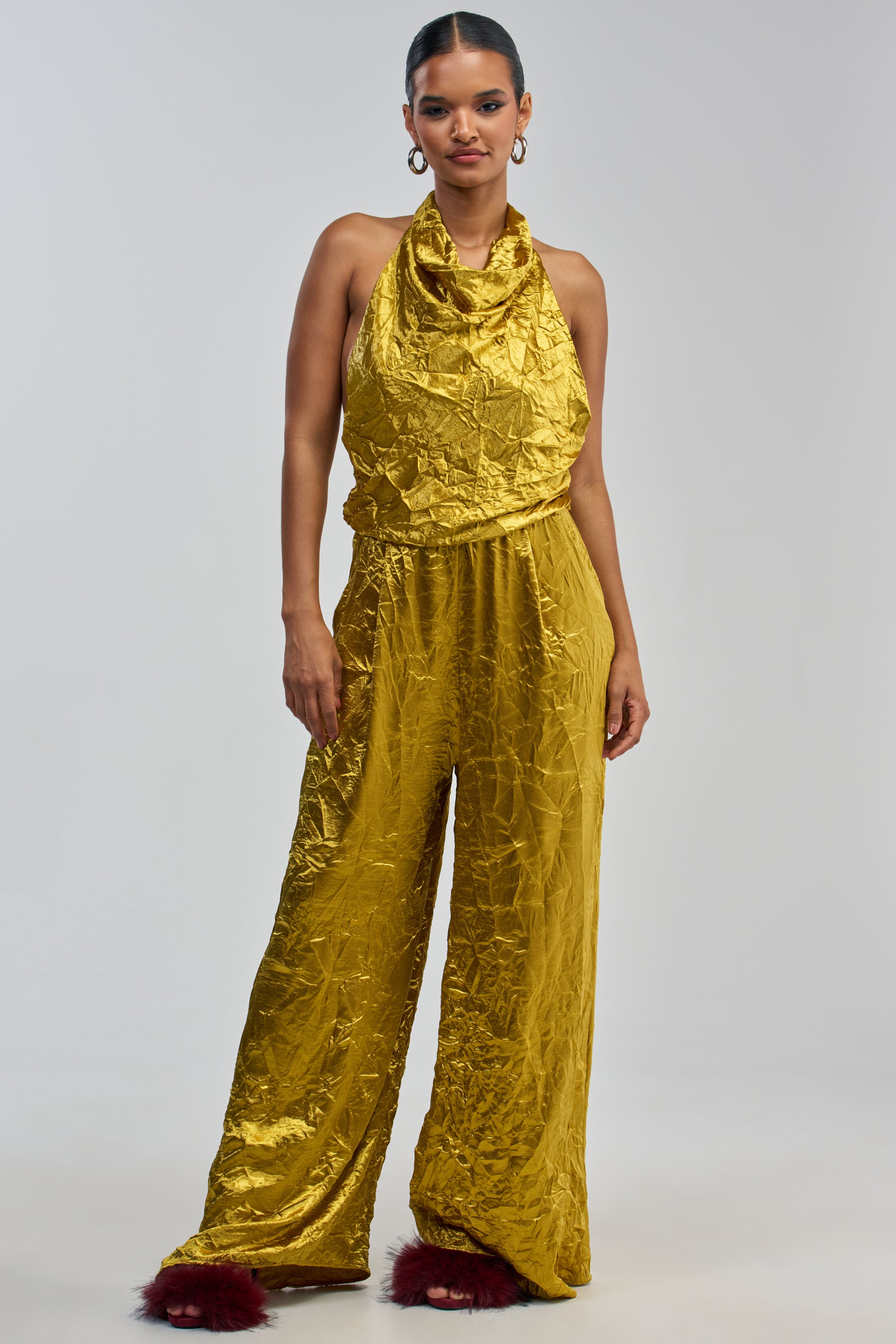 A woman models the CALLING ALL THE GIRLS SATIN JUMPSUIT IN GOLDEN YELLOW—a shiny, halter-style jumpsuit with wide legs—standing against a plain light gray background. Her hair is tied back, and she accessorizes with large hoop earrings and red furry slippers, creating an eye-catching outfit that’s perfect for elegant summer occasions.