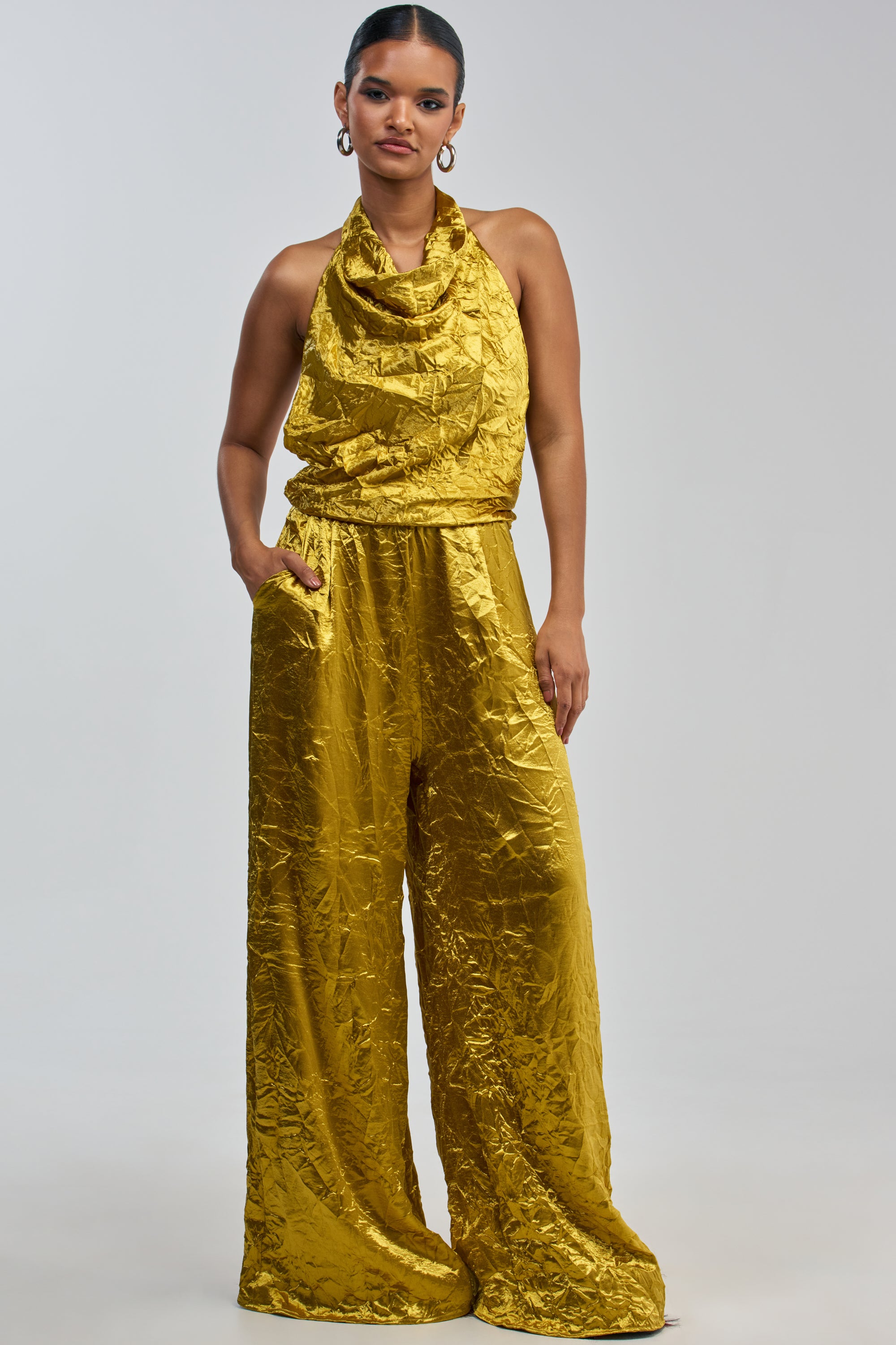 A woman models the CALLING ALL THE GIRLS SATIN JUMPSUIT IN GOLDEN YELLOW, featuring a halter neckline and wide-leg pants, striking a pose with one hand in her pocket against a simple light background—capturing an effortlessly chic take on elegant summer style.