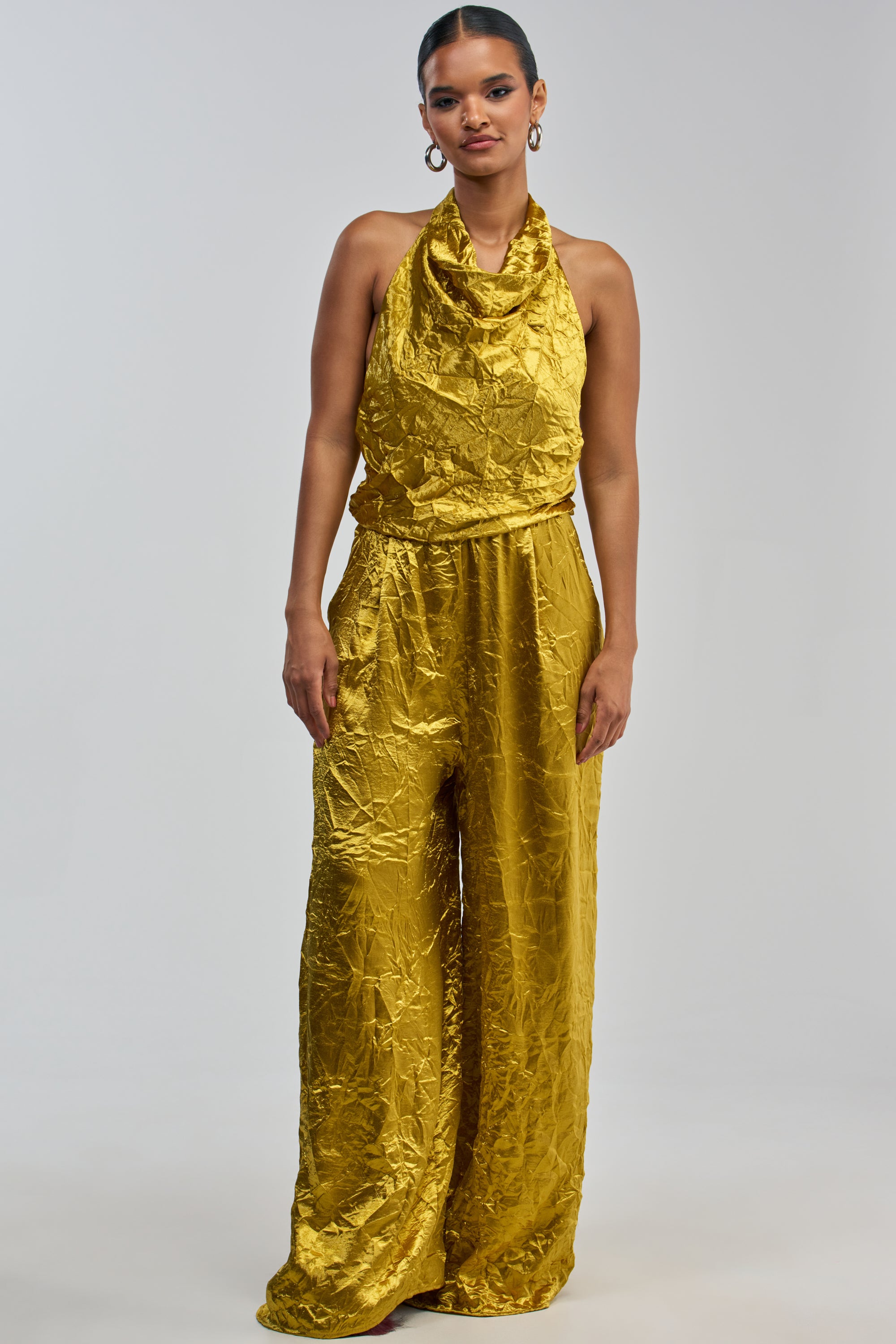 A woman stands against a plain light gray background wearing the CALLING ALL THE GIRLS SATIN JUMPSUIT IN GOLDEN YELLOW—an elegant summer piece featuring a halter neckline and wide-leg pants.