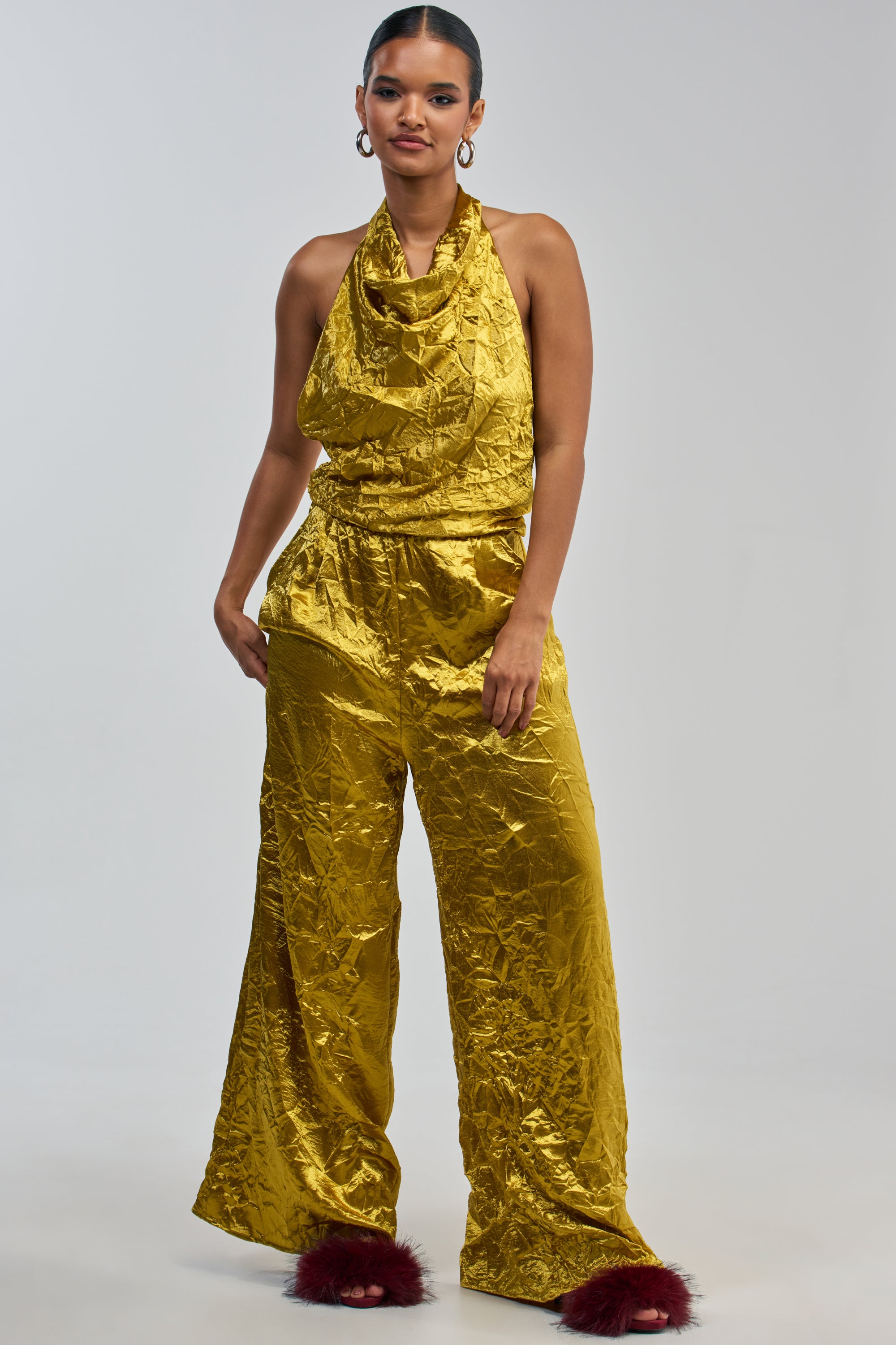 A woman stands confidently against a plain light gray background, wearing the CALLING ALL THE GIRLS SATIN JUMPSUIT IN GOLDEN YELLOW—a shiny gold halter-style jumpsuit with wide-leg pants reminiscent of an elegant summer ensemble—paired with burgundy fuzzy slippers.