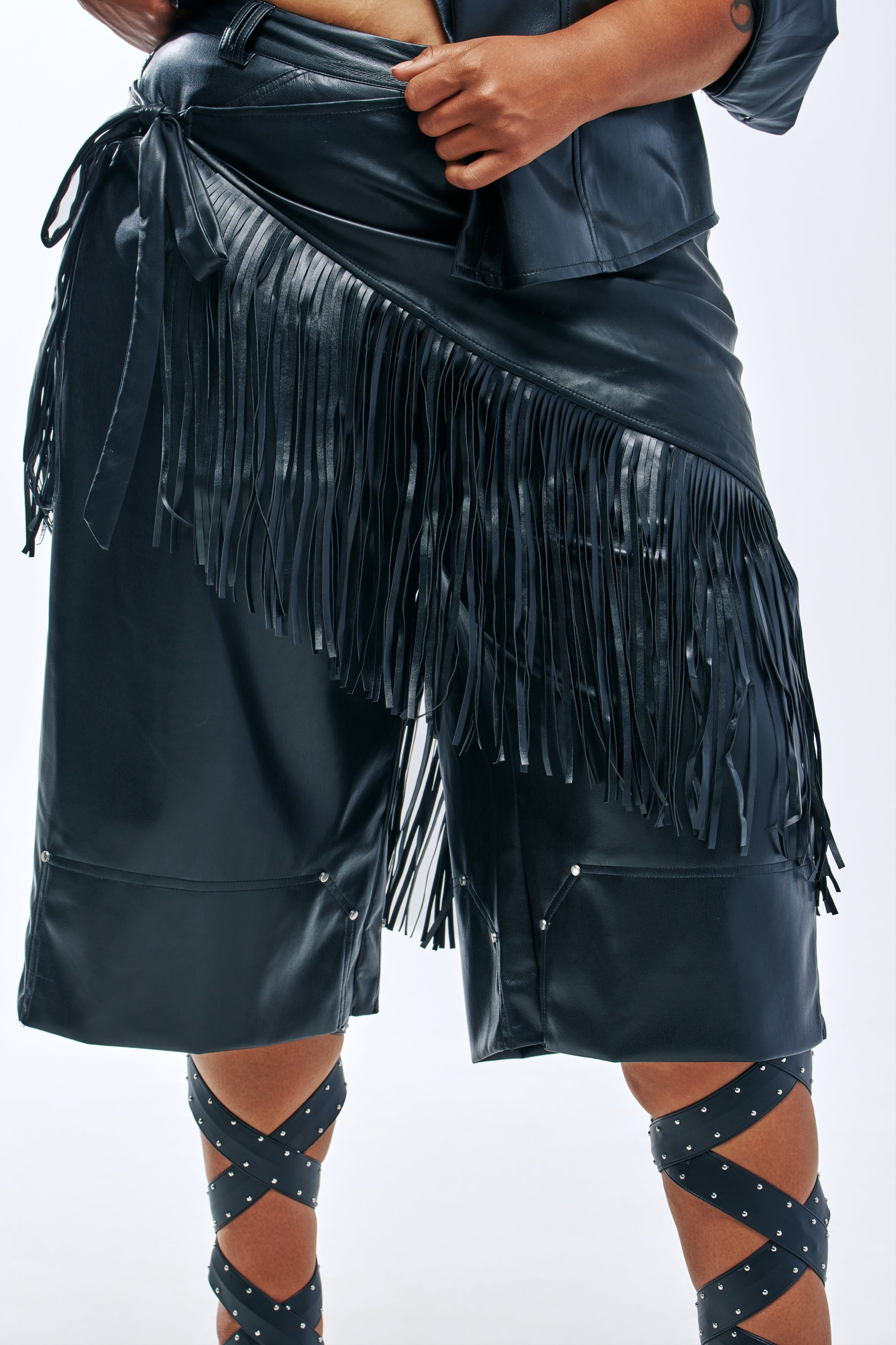 A black faux leather ALL THAT FRINGE TIE BELT with long fringe worn over black leather shorts.