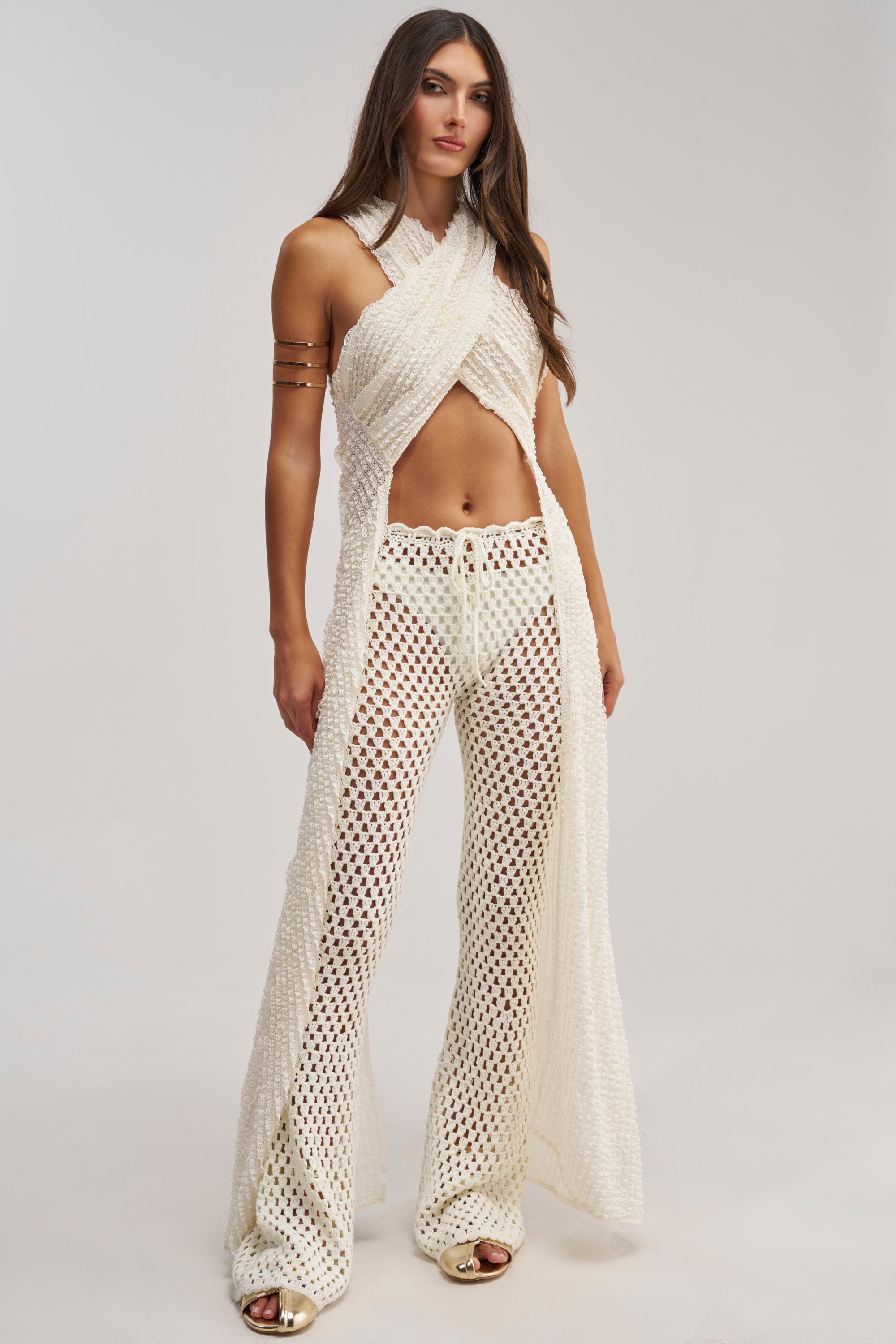 A woman stands against a plain background wearing the CIAO BELLA CRISS CROSS TOP in cream with matching wide-leg, see-through crochet pants featuring a drawstring waist. She has long hair and gold open-toe shoes.