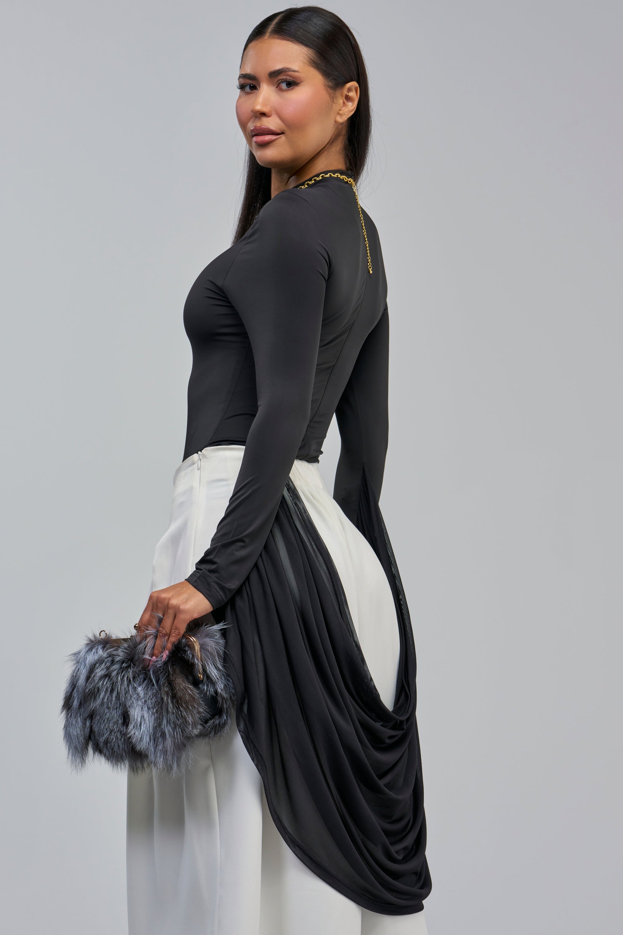 Wearing the ETHEREAL DRAPED SLEEVE BODYSUIT, this woman pairs its elegant, fitted black silhouette with white pants and a fur clutch. Against a plain gray background, she glances over her shoulder—an effortlessly chic look perfect for a girls’ night out.
