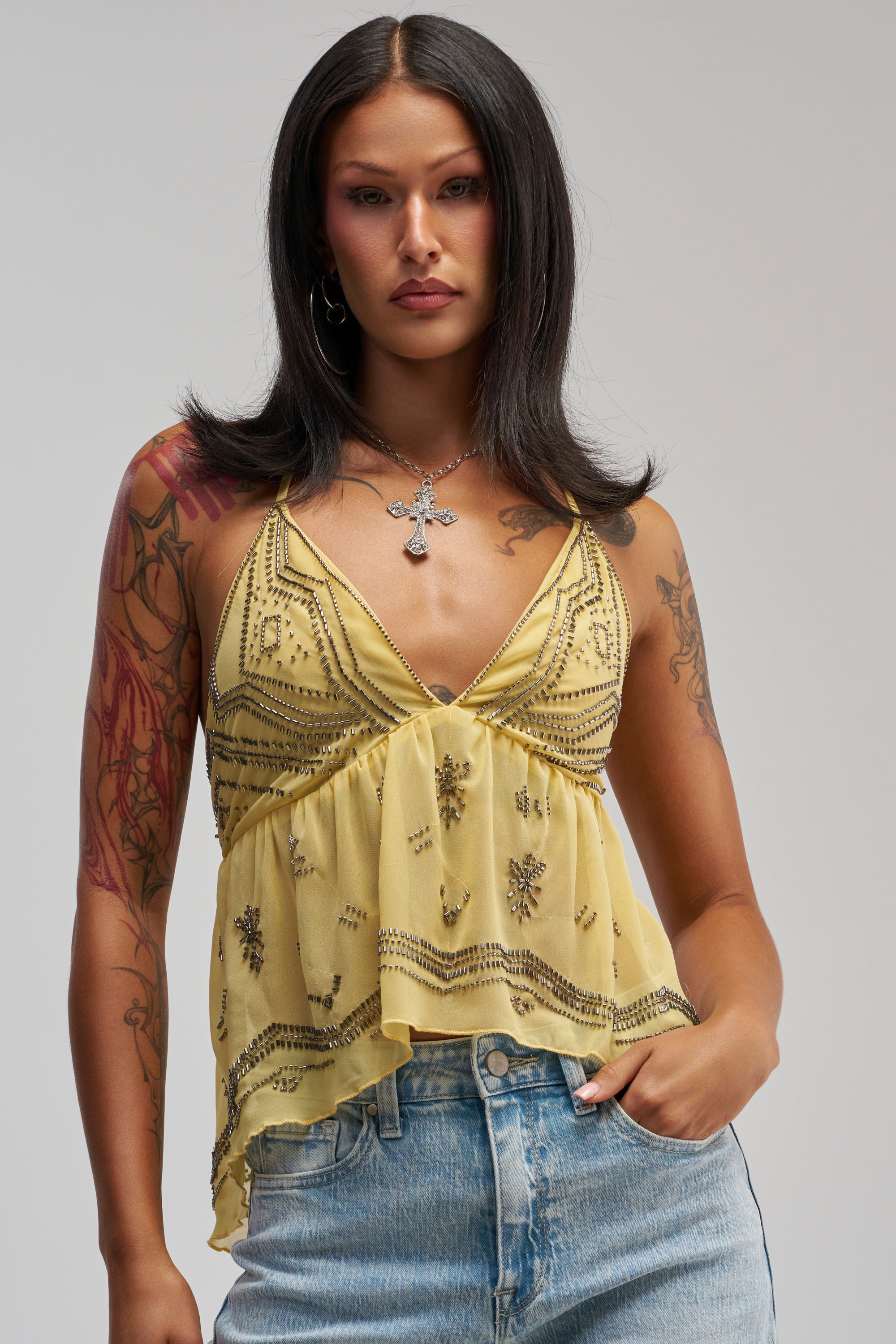 A woman with long dark hair wears the 90′S BEADED CHIFFON TOP IN YELLOW paired with blue jeans, effortlessly channeling festival styling. She has tattoos on her arms, a silver cross necklace, and is standing against a plain gray background.