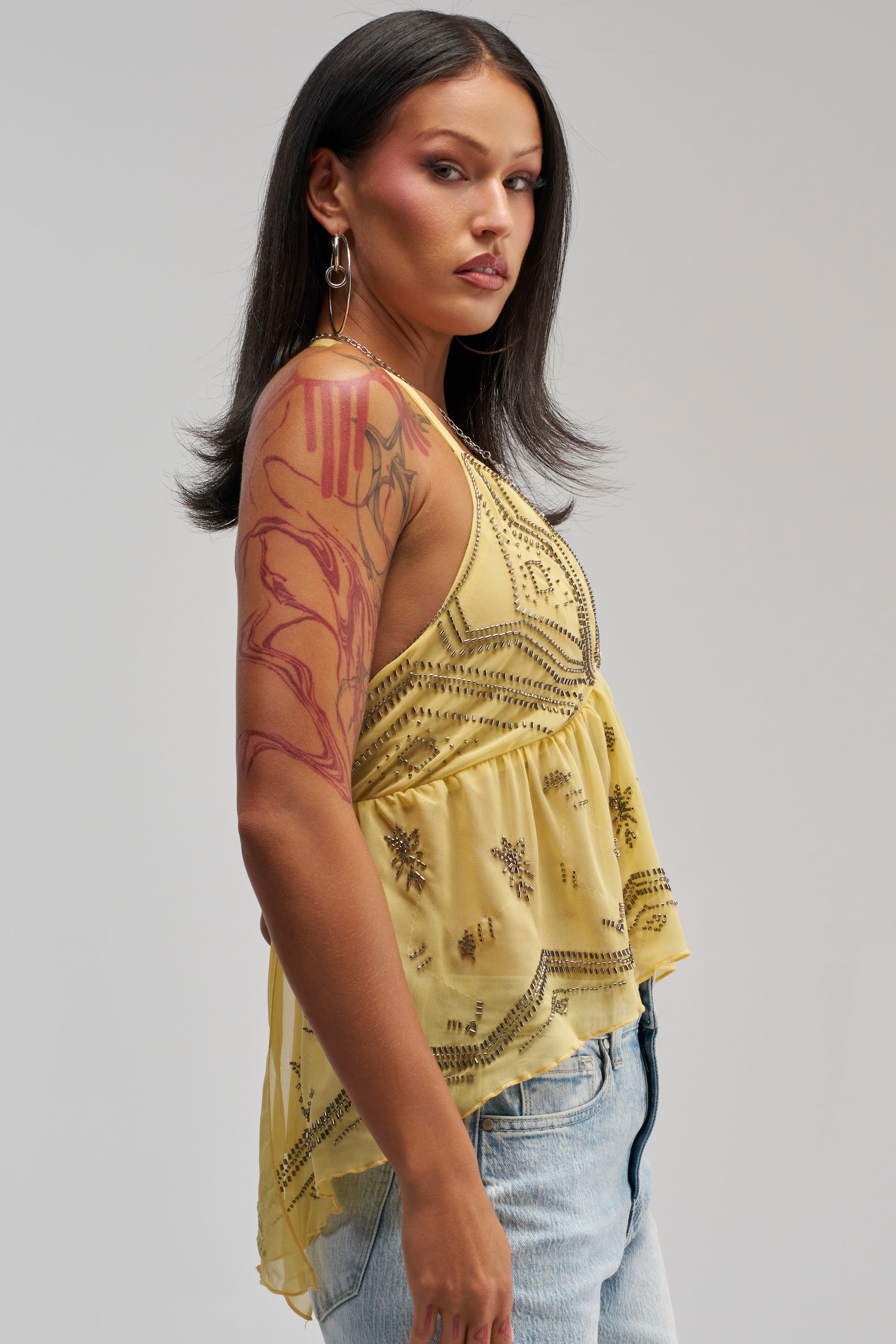 A woman with long dark hair, pink eyeshadow, and a large arm tattoo models the 90′S BEADED CHIFFON TOP IN YELLOW paired with light blue jeans, standing sideways against a plain grey background—an effortlessly chic look that’s perfect for festival styling or summer wear.