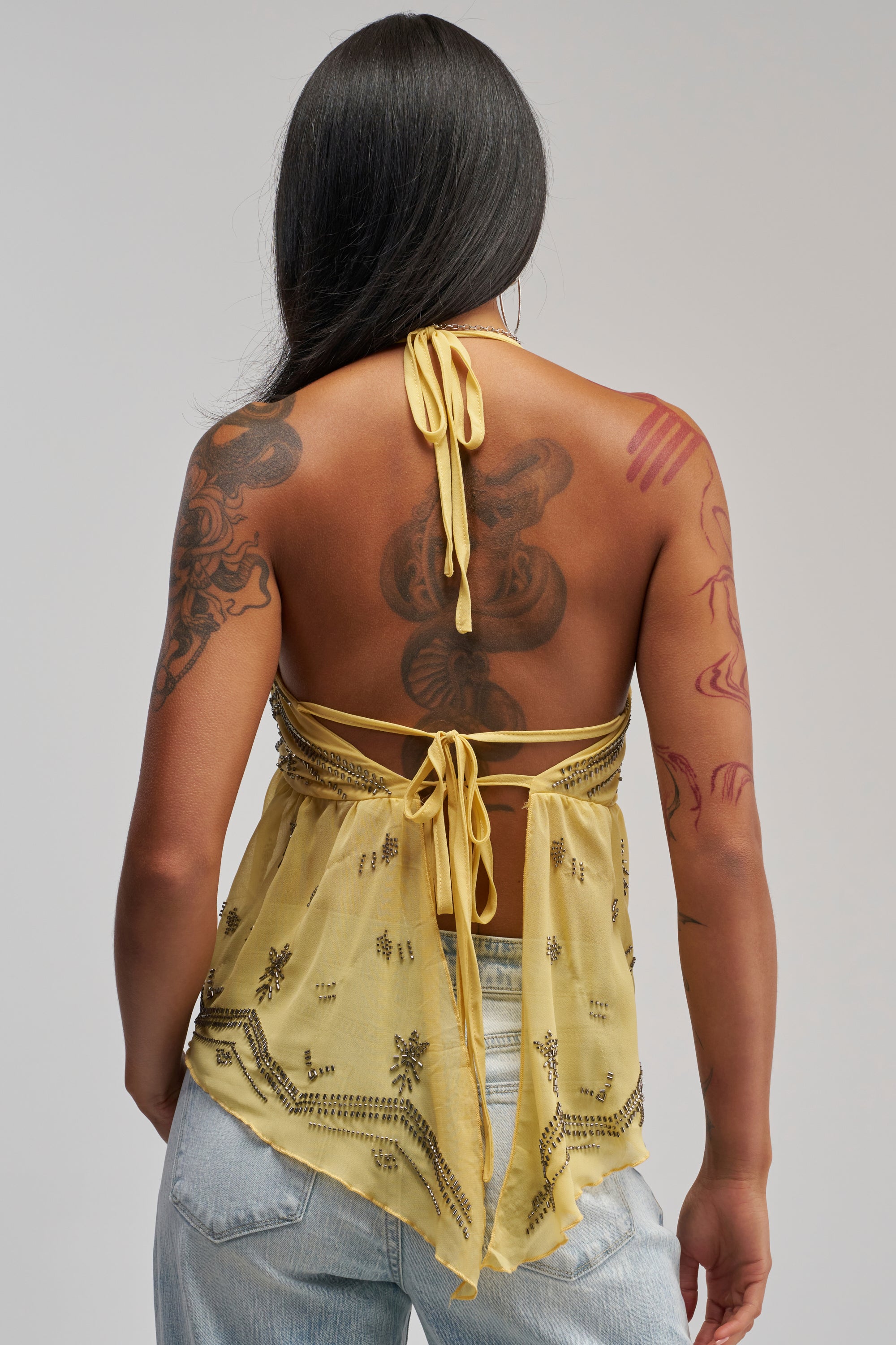 A person with long dark hair and tattoos on their back and arms faces away from the camera, wearing light blue jeans paired with the 90′S BEADED CHIFFON TOP IN YELLOW—an effortless choice for summer wear and festival styling.