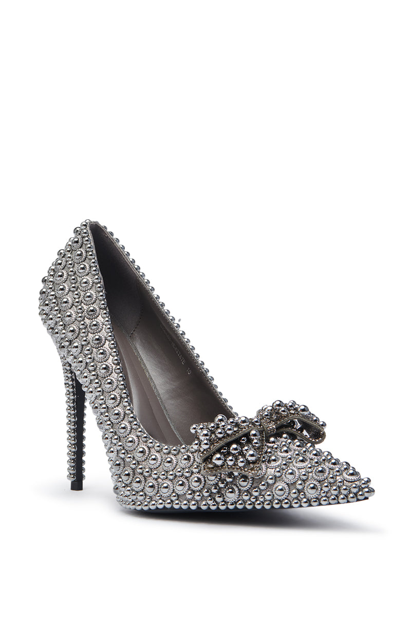 The AZALEA WANG HEMITATE STONE EMBELLISHED PUMP is a faux leather, high-heeled, pointed-toe shoe adorned with silver rhinestones and a matching bow on the front, featuring a sleek stiletto heel against a plain white background.