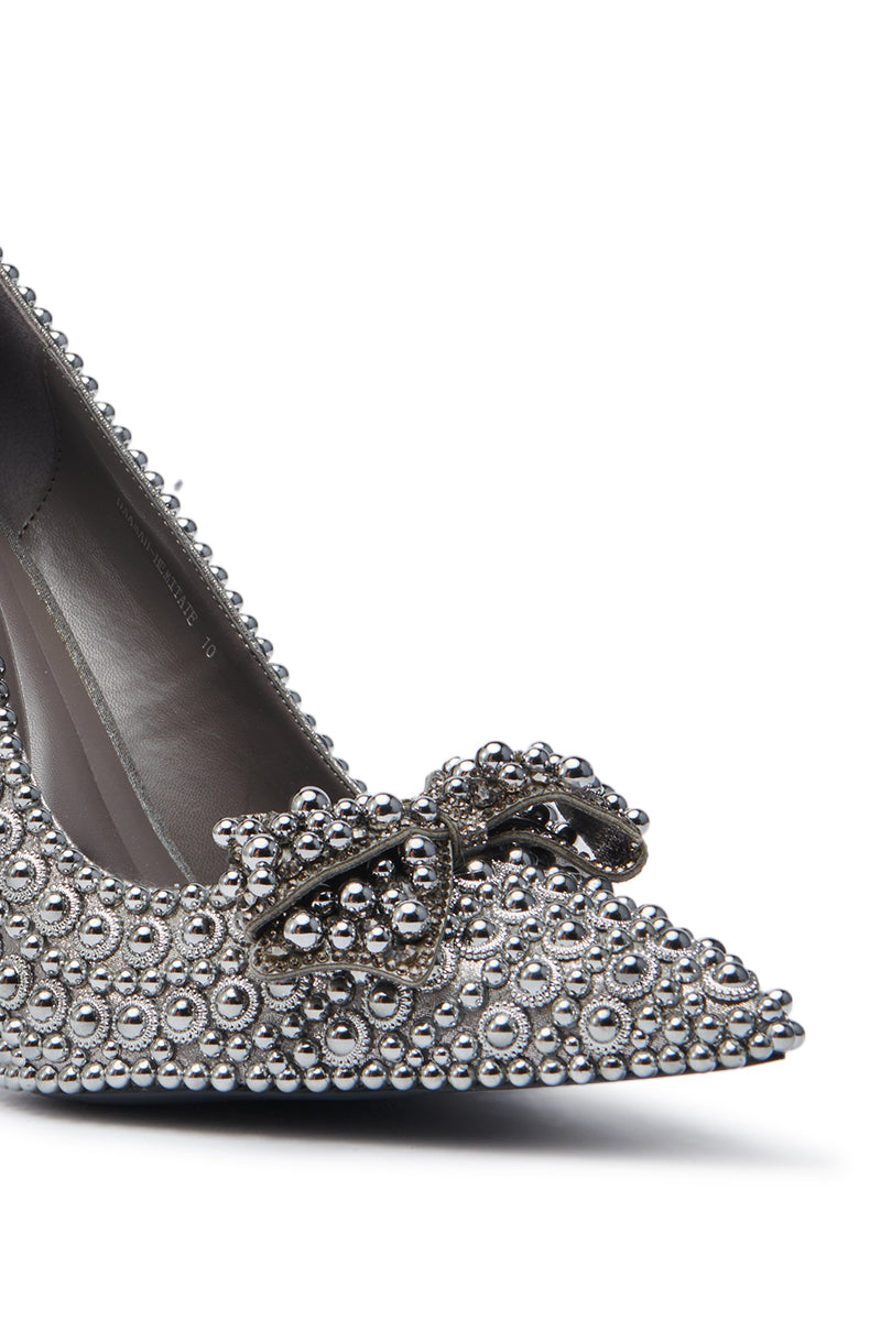 The AZALEA WANG HEMITATE STONE EMBELLISHED PUMP is a faux leather pointed-toe stiletto decorated with silver studs, rhinestones, and a bow on the toe, shown close-up against a white background.