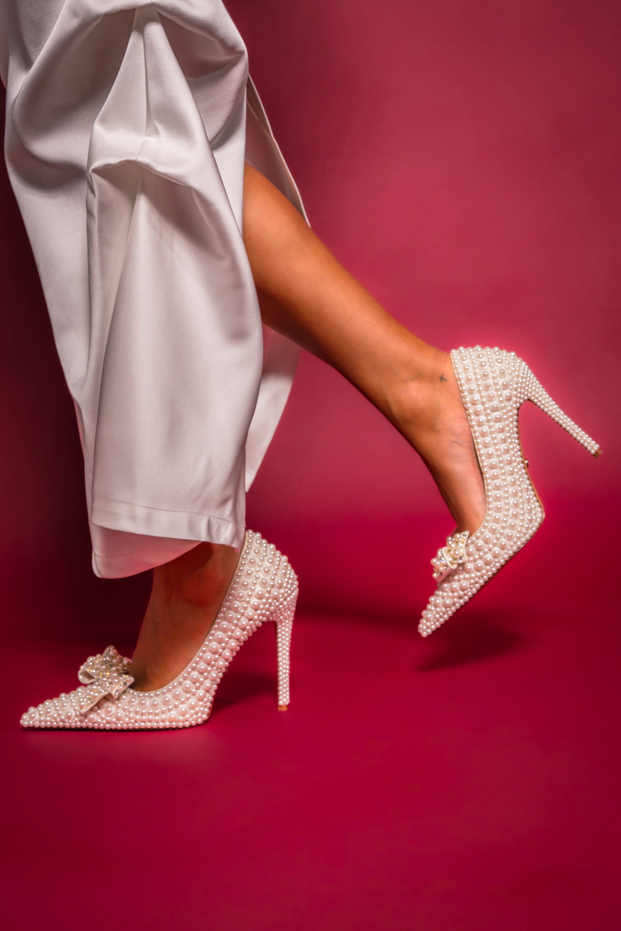 A person poses against a pink background wearing white satin pants and the AZALEA WANG NAAMAH WHITE PEARL EMBELLISHED PUMP, featuring elegant stiletto heels adorned with pearls and bows.