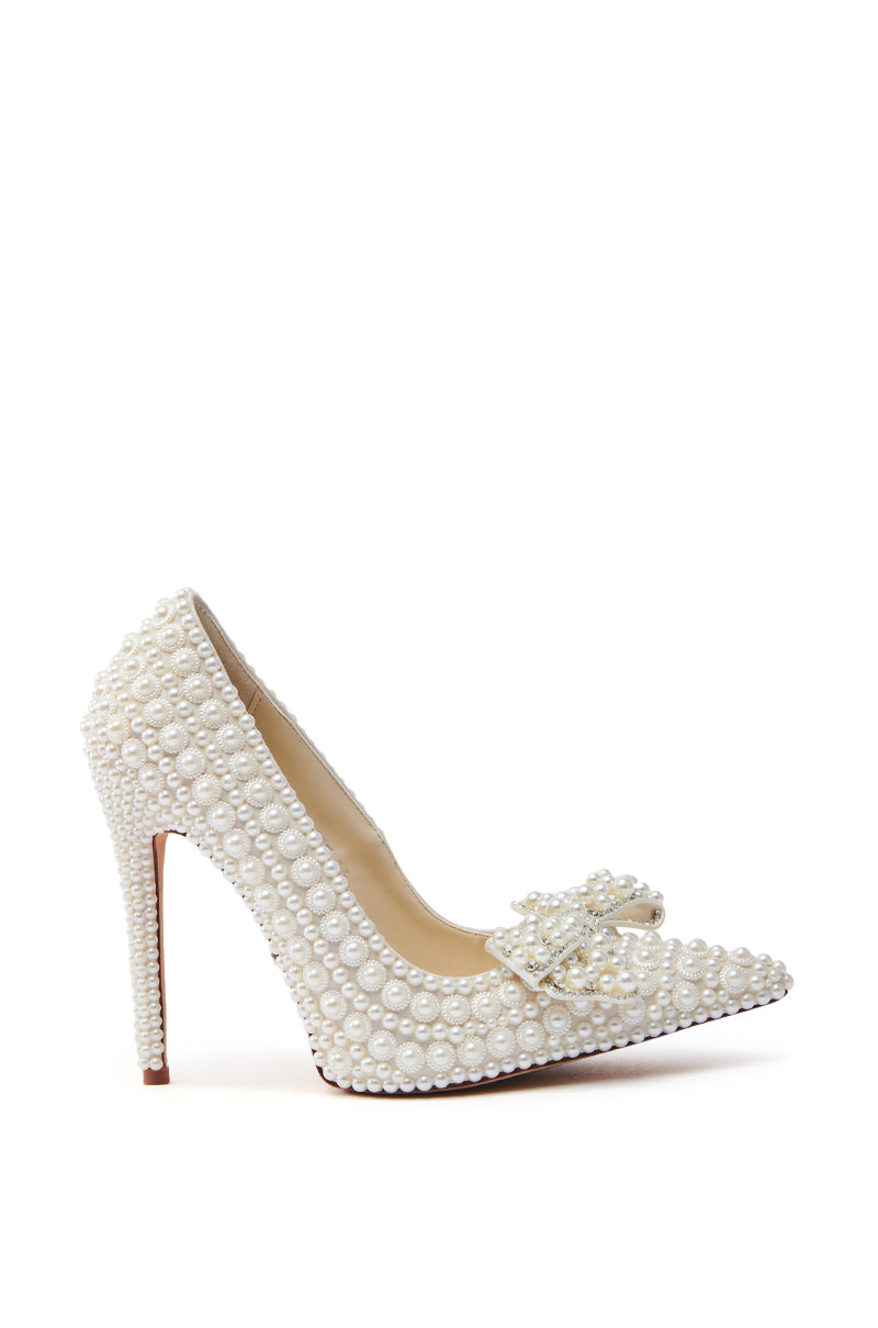 The AZALEA WANG NAAMAH WHITE PEARL EMBELLISHED PUMP is a white, pointed-toe high heel made from vegan leather and adorned with pearl embellishments. It features a pearl-embellished bow on the front.