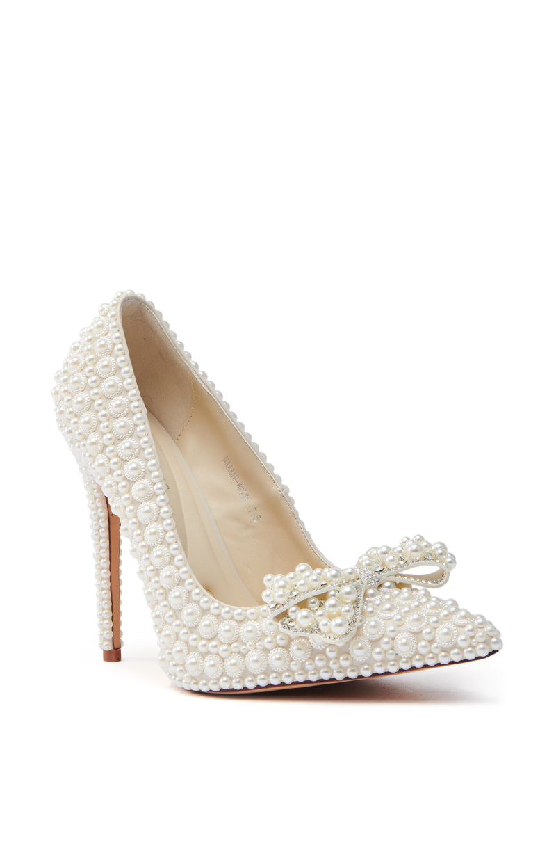 The AZALEA WANG NAAMAH WHITE PEARL EMBELLISHED PUMP is a sleek white high heel crafted from vegan leather, featuring a pointed toe, decorative bow, and stiletto heel. Adorned with pearl embellishments, it exudes elegance and luxury.