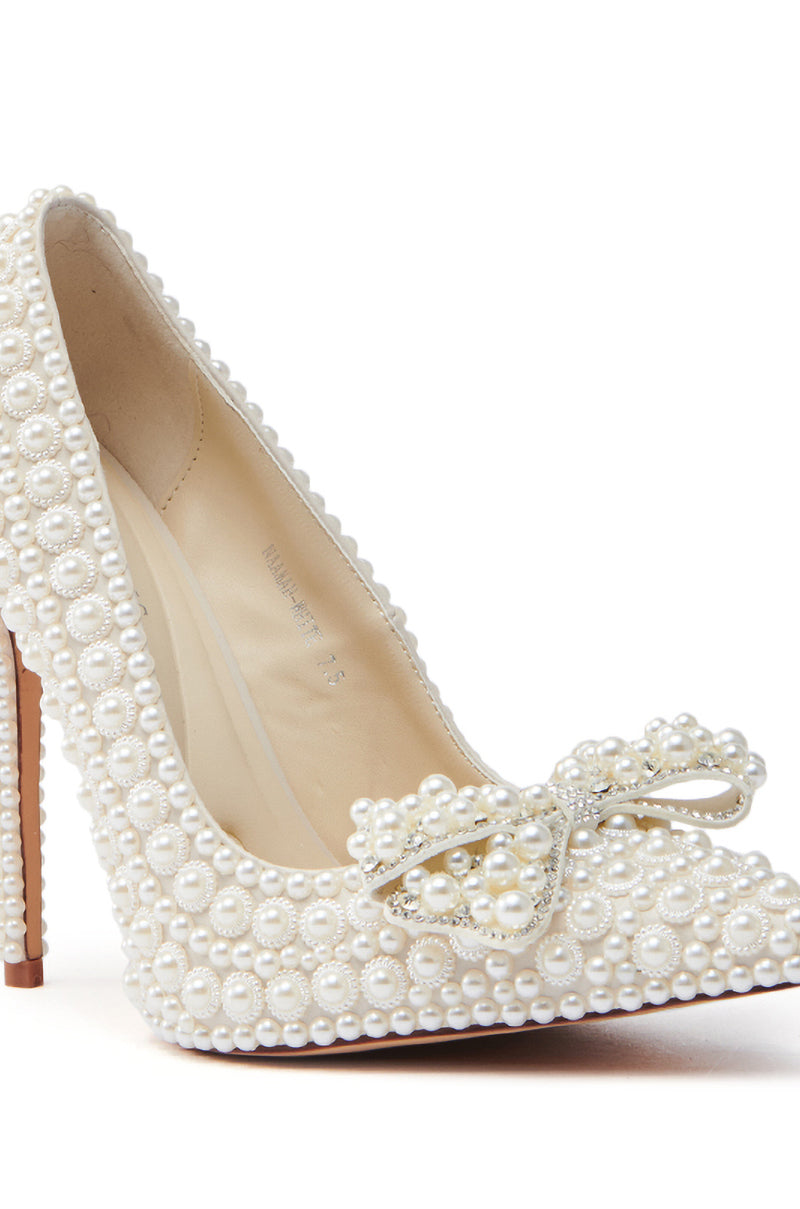 The AZALEA WANG NAAMAH WHITE PEARL EMBELLISHED PUMP is a sophisticated high-heeled shoe. Made from vegan leather, it showcases a pointed toe, ornate pearl bow, and chic stiletto heel, all adorned with white pearls, complemented by a light tan sole.