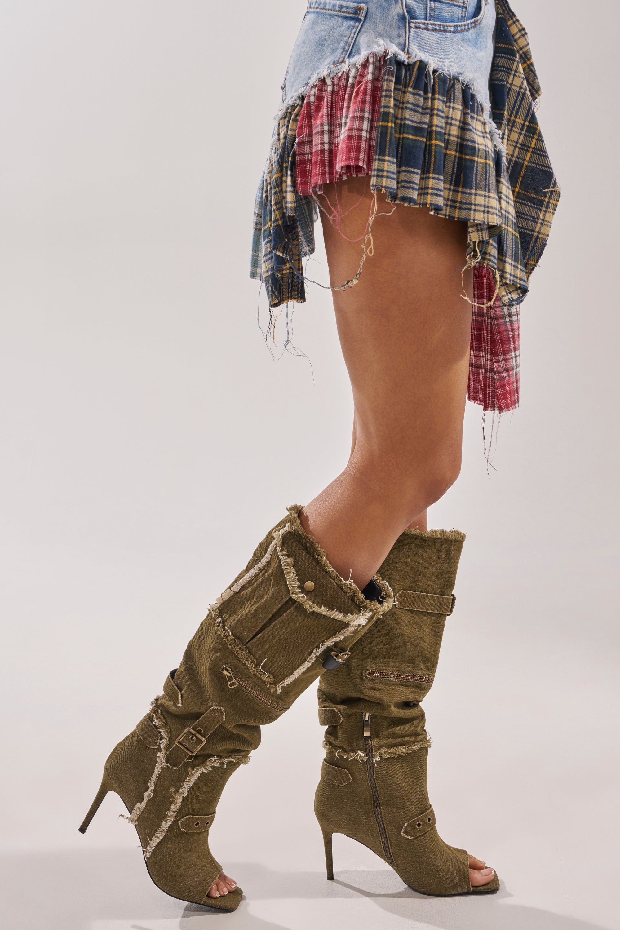 A woman in a frayed denim and plaid skirt stands in profile, showing off the AKIRA GOLD NADAL OLIVE CANVAS OPEN TOE STILETTO BOOTIE—chic festival footwear with stiletto heels and bold details, set against a plain background. 2