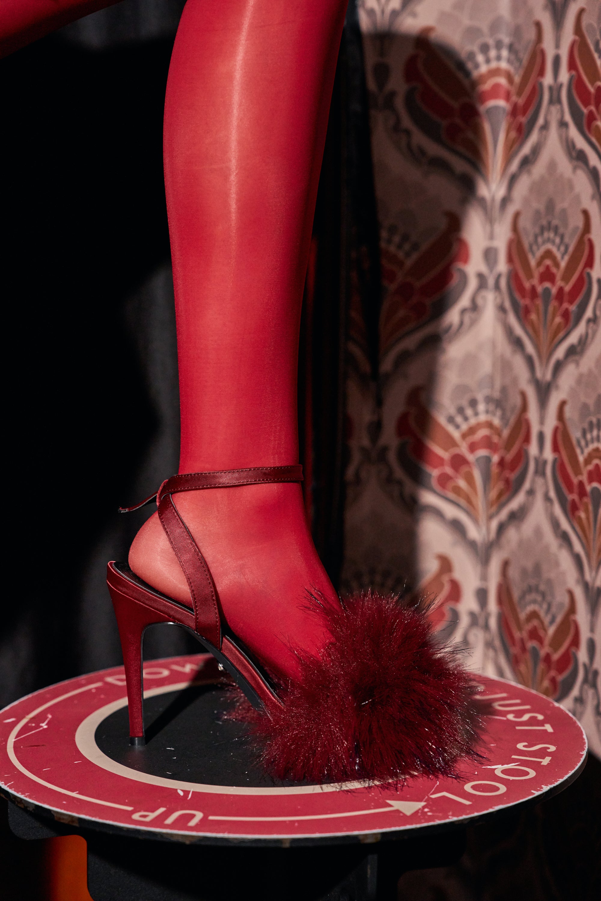 A leg in red tights wearing an AZALEA WANG NADIA BURGUNDY FUR EMBELLISHED STILETTO SANDAL featuring a burgundy satin heel and a large faux fur embellishment on the front strap.