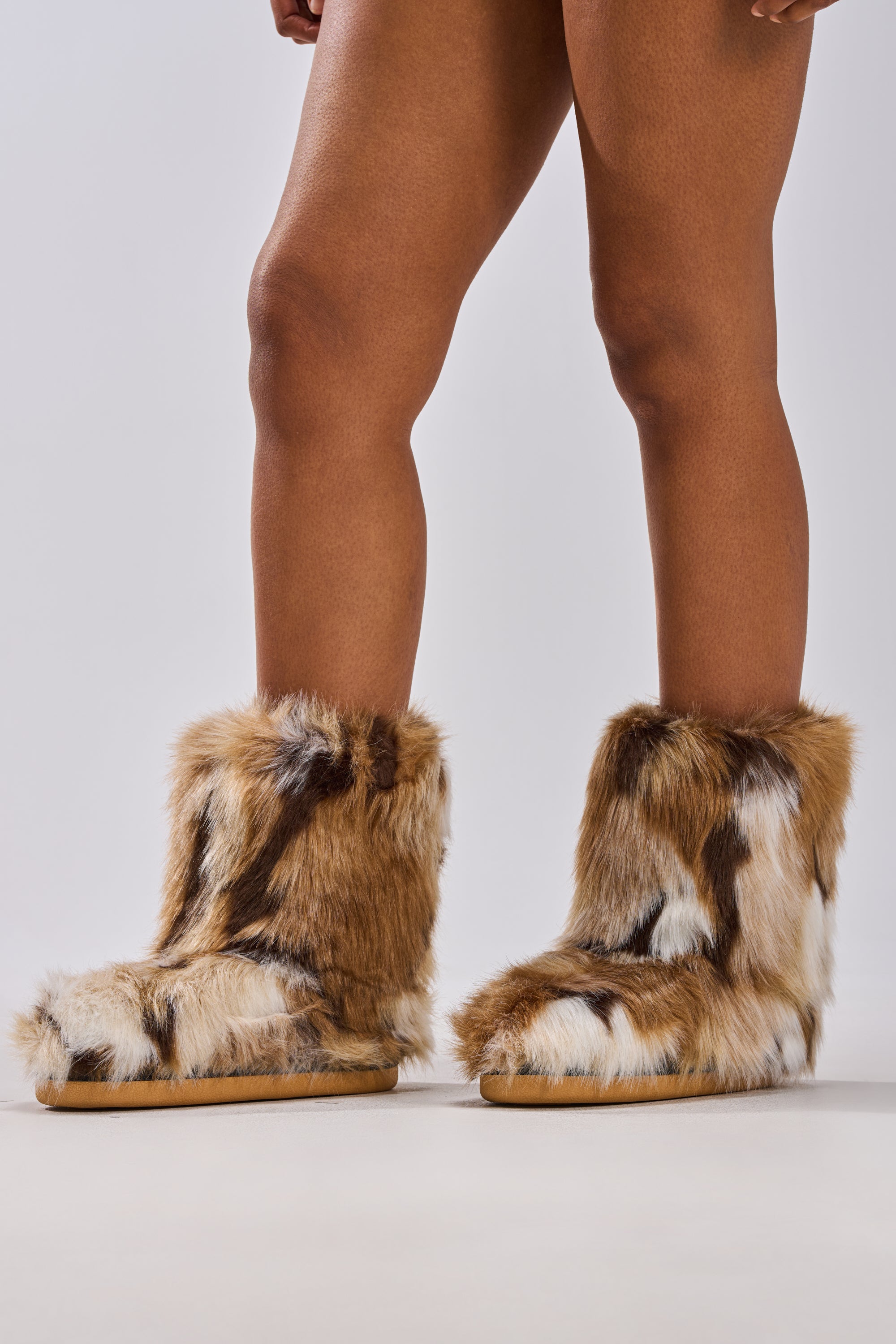 A person stands on tiptoe, showcasing bare legs and the AZALEA WANG NADIANNA BROWN FUR FLAT BOOTIE, which reaches mid-calf, set against a plain light grey background.