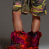 A person showcases a striking blend of winter fashion with colorful faux fur boots and camouflage shorts against a neutral background. The vibrant shades of the AZALEA WANG NADIANNA ORANGE BOOTIE add a bold contrast to the patterned shorts, creating a daring and stylish look.