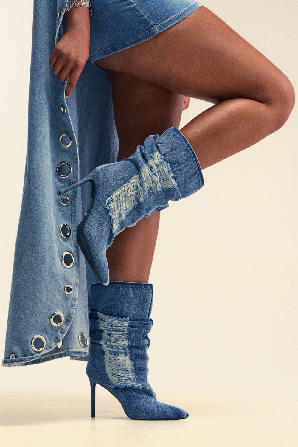 A person wearing a long denim skirt with grommet details and the AZALEA WANG NAELIA DENIM STILETTO BOOTIE—knee-high, pointed-toe, high-heeled denim boots featuring distressed patches—stands with one leg crossed over the other, perfectly embodying streetwear festival boots style.