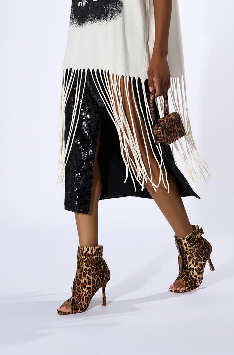 A person wearing a white fringe dress and black sequin skirt is elegantly walking in stiletto heels featuring the AZALEA WANG NAILY CHEETAH BUTTON UP BOOTIE. They are holding a small matching leopard print handbag against a plain white background. 1