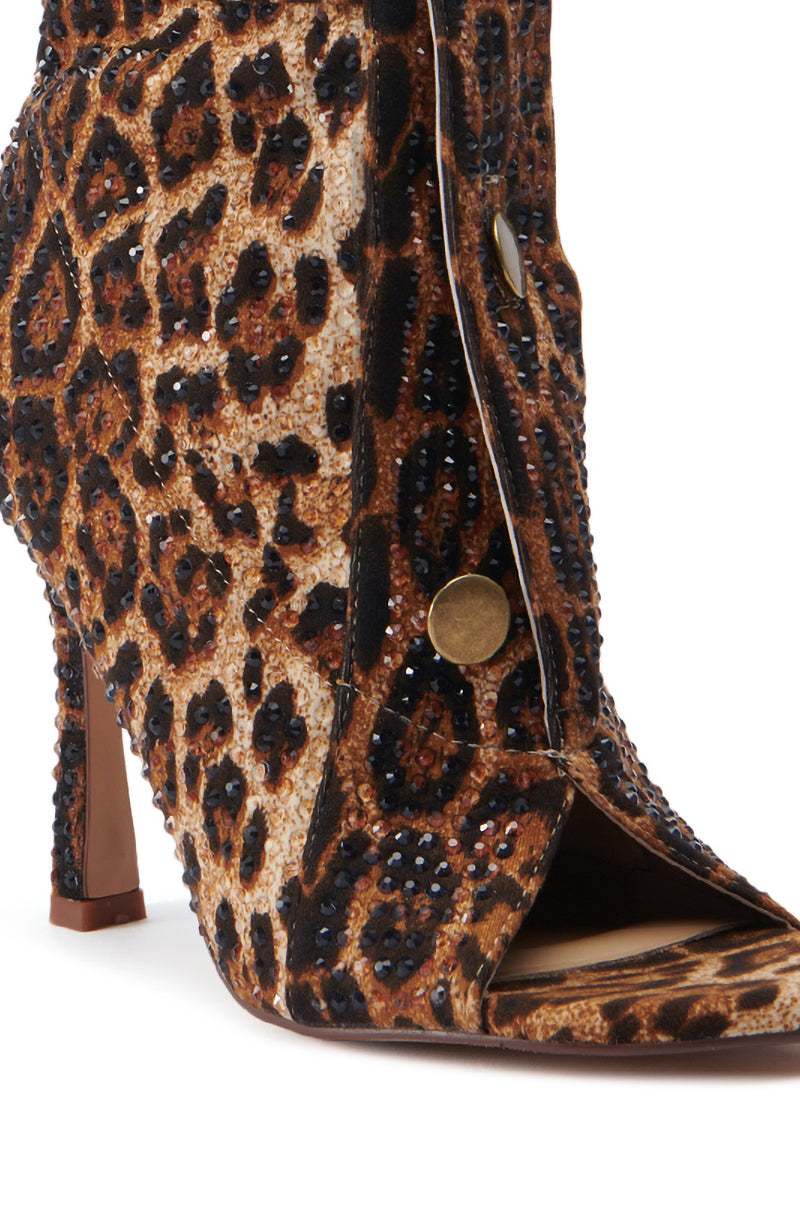 A close-up of the AZALEA WANG NAILY CHEETAH BUTTON UP BOOTIE. This high-heeled shoe boasts a cheetah print pattern, an open toe adorned with small, dark rhinestones, and a series of metallic button details along its side. 5