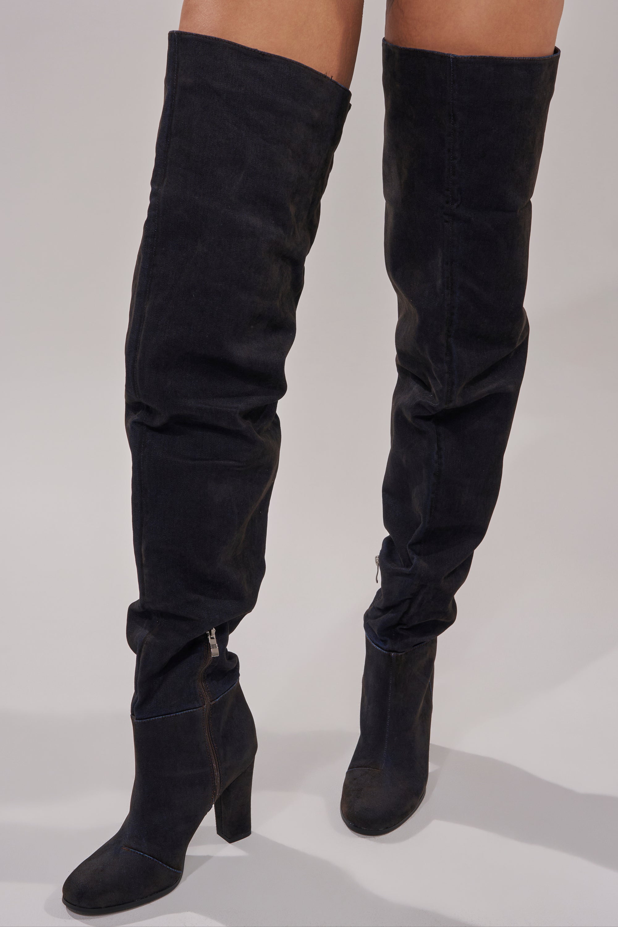 A person wears the AZALEA WANG NANETTE BLACK DENIM SLOUCHY BLACK ROUND TOE CHUNKY BOOT with a slightly slouchy fit, side zippers, and chunky heels, standing on a light gray surface for an edgy look.