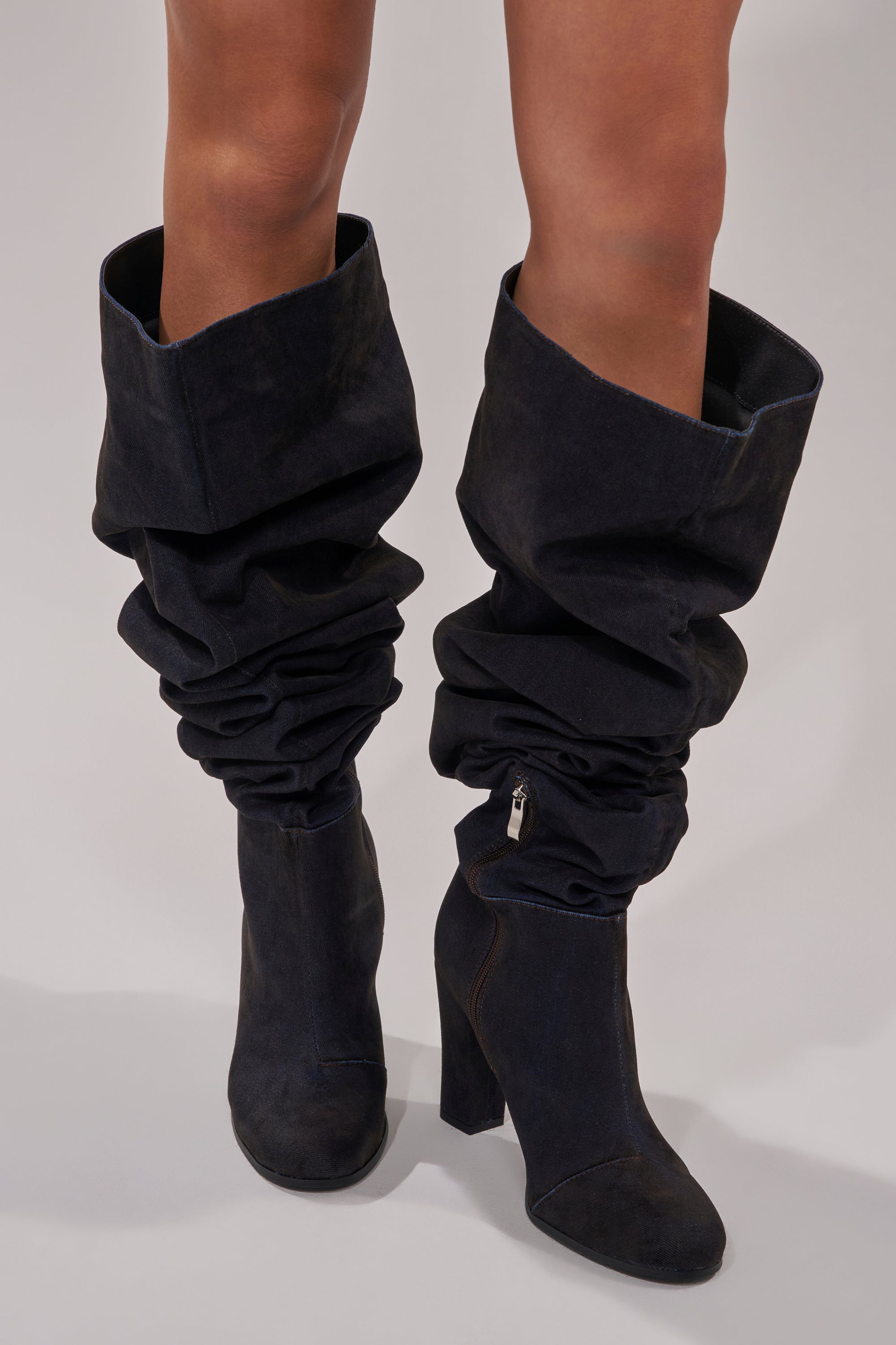 A person wearing AZALEA WANG NANETTE Black Denim Slouchy Round Toe Chunky Boots that reach just below the knees, standing on a light gray surface.