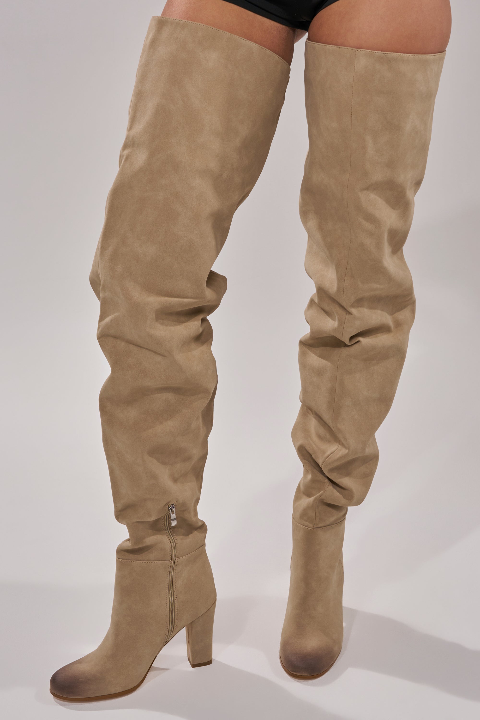 Someone models the AZALEA WANG NANETTE TAUPE SLOUCHY BOOT, featuring tan faux suede, chunky high heels, and side zippers, while standing against a plain light background.