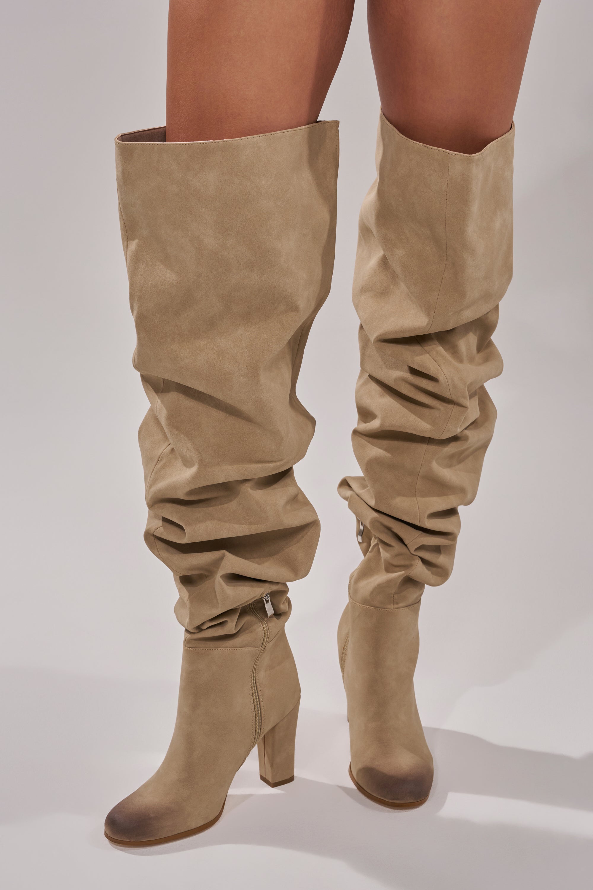 A person stands against a neutral background wearing the AZALEA WANG NANETTE TAUPE SLOUCHY BOOT, a tan, thigh-high boot with a chunky heel.