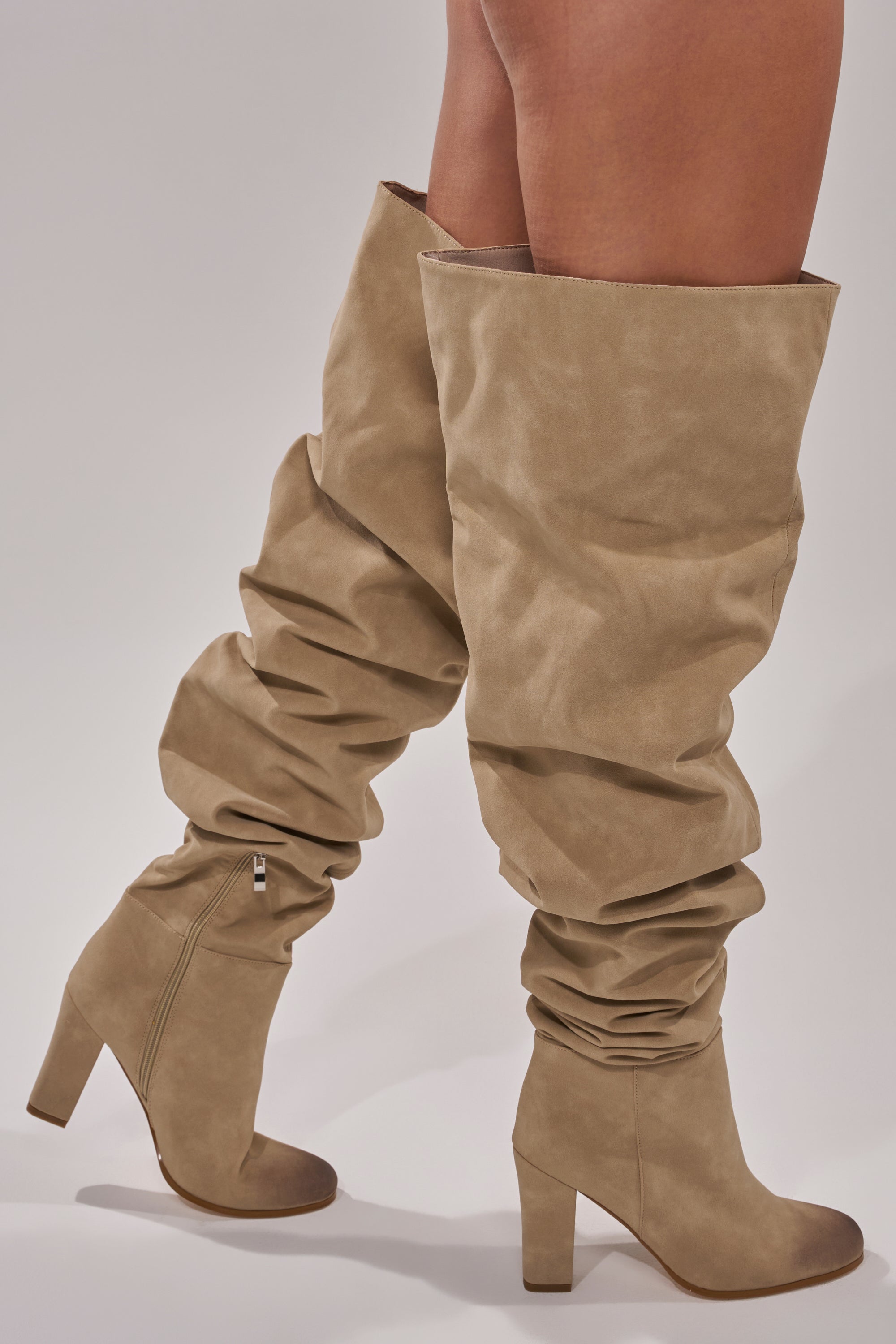 Someone wearing the AZALEA WANG NANETTE TAUPE SLOUCHY BOOT, featuring tall thigh-high taupe faux suede, side zippers, and a chunky heel, stands against a plain light background.