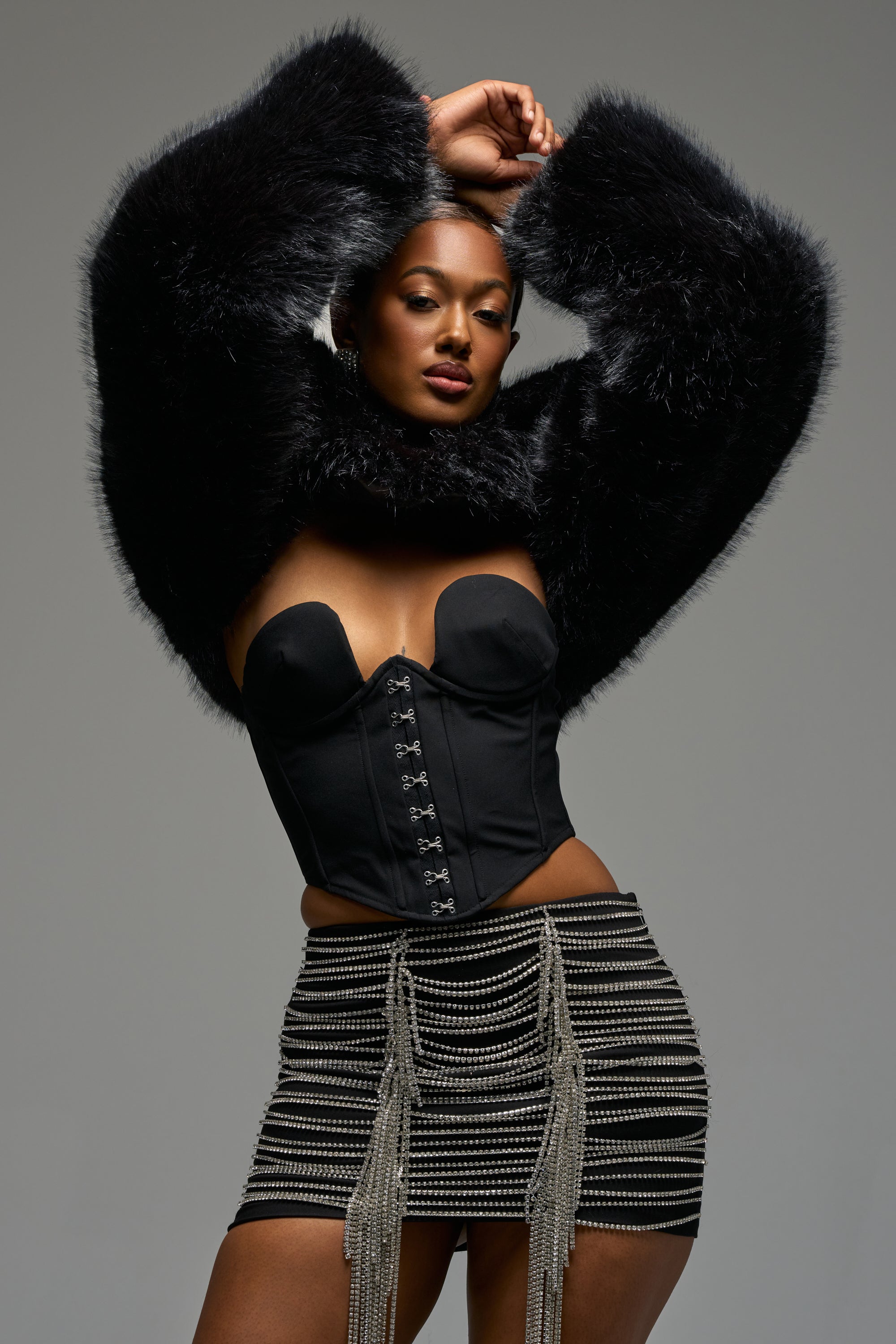 Wearing the LEILA ULTRA CROP FAUX FUR IN BLACK, a woman poses in a strapless black corset and short skirt with metallic chain details against a plain gray background.