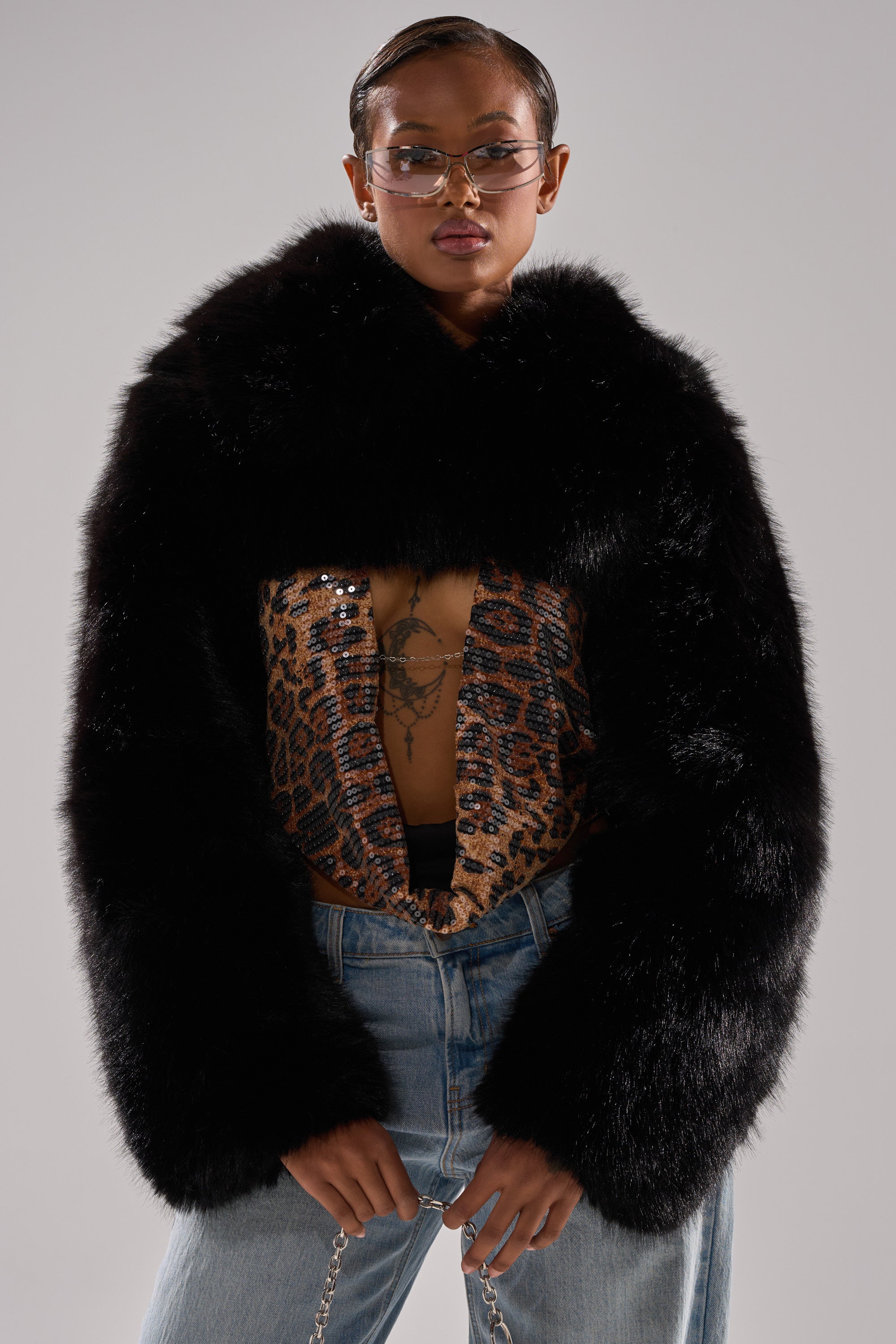 Person wearing the LEILA ULTRA CROP FAUX FUR IN BLACK over a sequined animal print top, with blue jeans, clear glasses, and silver jewelry, standing against a neutral background.