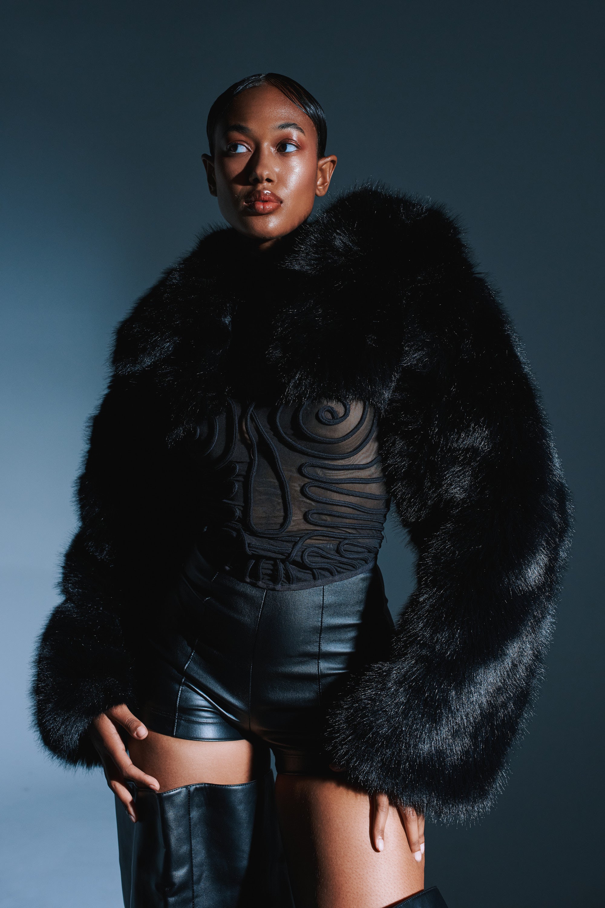 A stylish woman poses confidently in the LEILA ULTRA CROP FAUX FUR IN BLACK jacket, paired with a sheer black patterned top, high-waisted leather shorts, and thigh-high boots against a dark background.