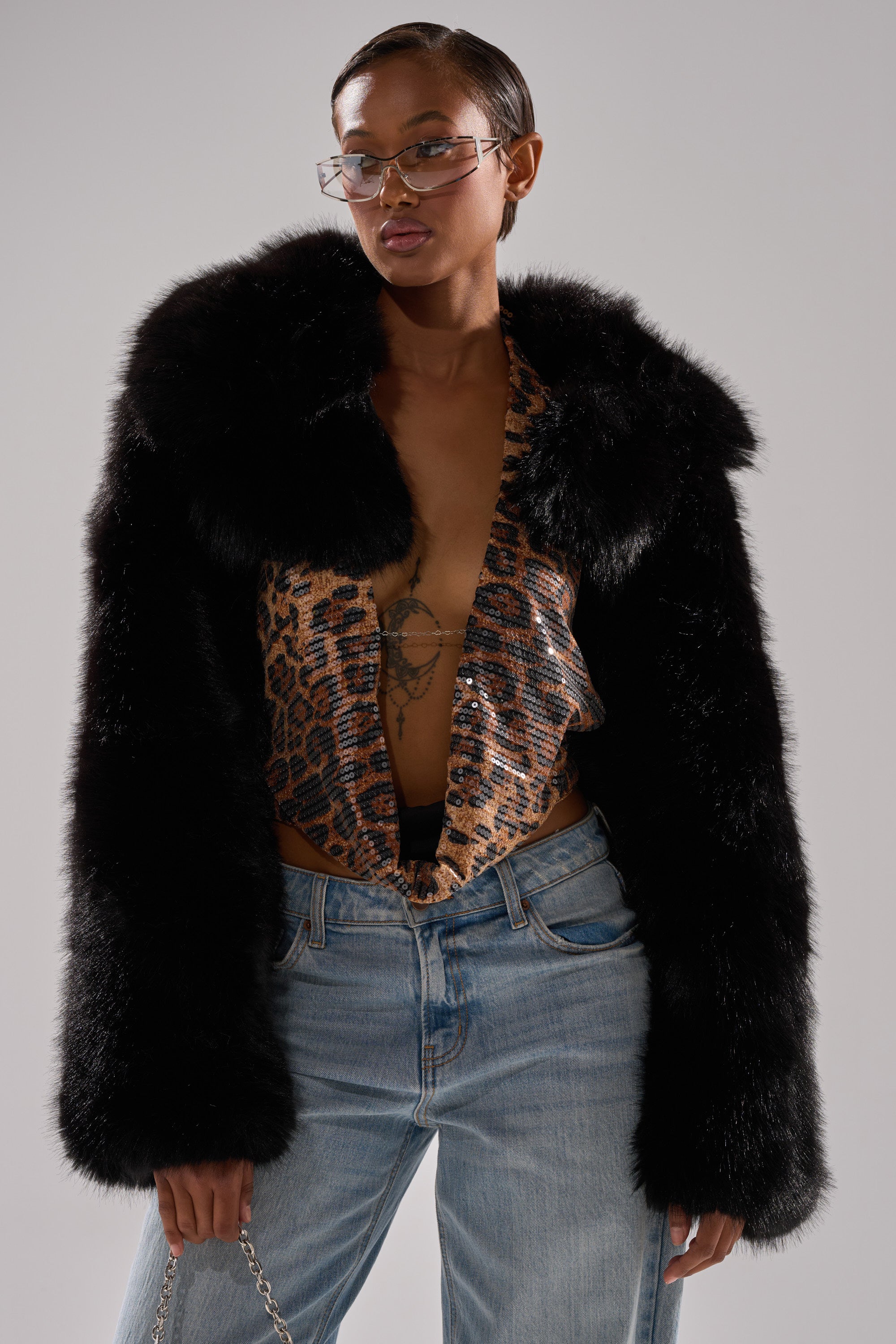 A person in wire-frame glasses, the LEILA ULTRA CROP FAUX FUR IN BLACK jacket, a leopard print sequined top, and light blue jeans stands with one hand in their pocket against a neutral background.