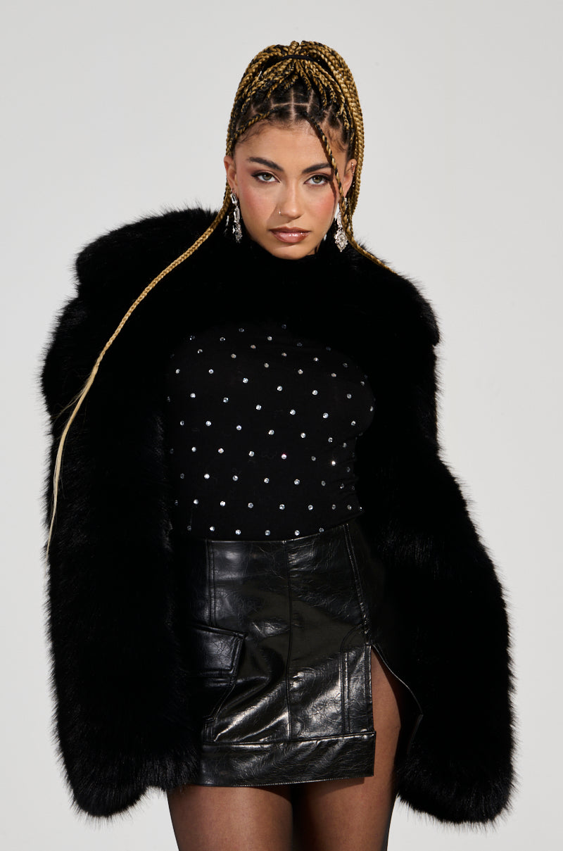 A person with braided hair is sporting the LEILA ULTRA CROP FAUX FUR IN BLACK over a sleek black leather skirt and an ultra crop black top adorned with silver dots. Bold earrings enhance their confident expression.