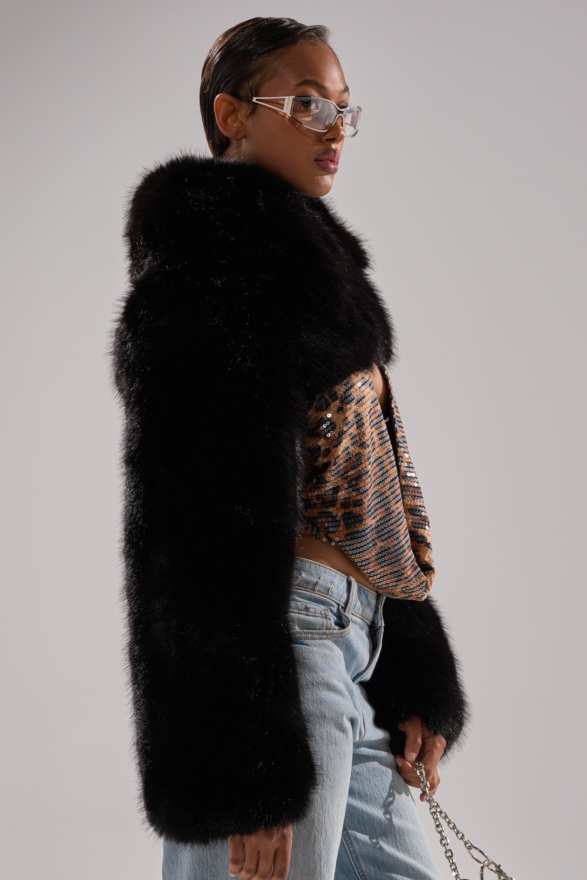 A person in clear glasses wears the LEILA ULTRA CROP FAUX FUR IN BLACK over an animal print top and light blue jeans, standing in profile against a plain background and looking confidently ahead.