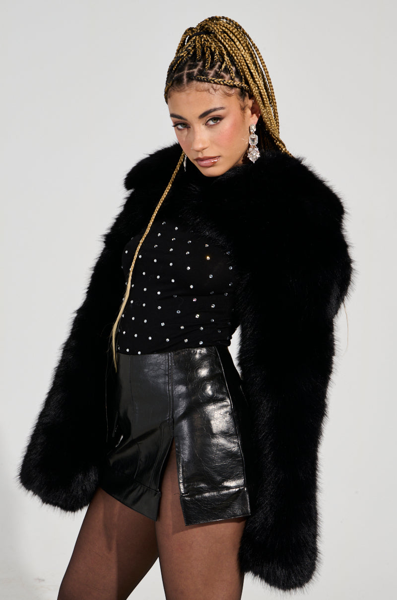 A person with braided hair is wearing the LEILA Ultra Crop Faux Fur in black, paired with a studded ultra crop black top and a sleek black leather skirt. They are striking a pose against a plain white background.